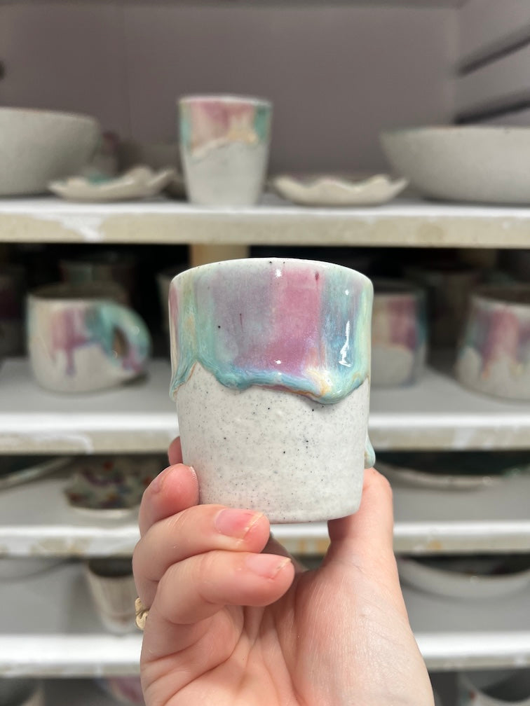 A hand holding a freshly fired cup in pink and turquoise glaze in front of a loaded kiln
