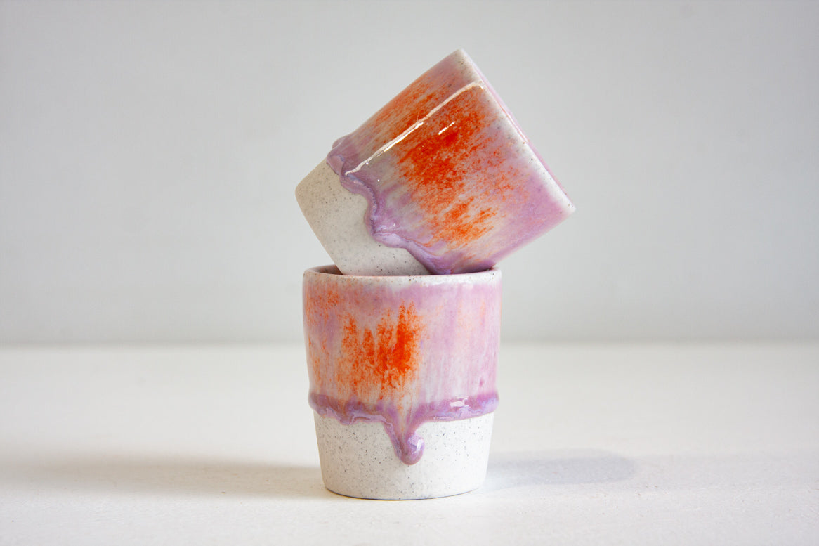 Two handmade ceramic espresso cups stacked at an angle, showing lilac and orange glaze with purple drips over speckled white stoneware