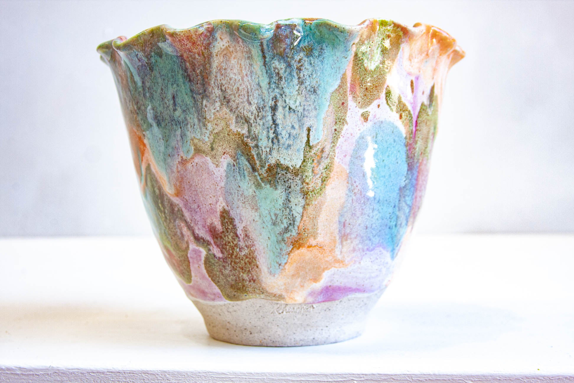A handmade ceramic frill vessel seen from the front, purple, green, peach and blue running down the flared sides to a bare white clay foot.