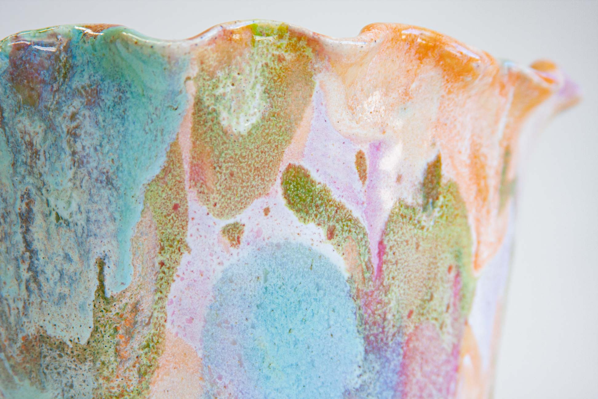 Close-up of a handmade ceramic frill vessel, blue and green pooling in soft patches across peach and lilac below the frilled rim.