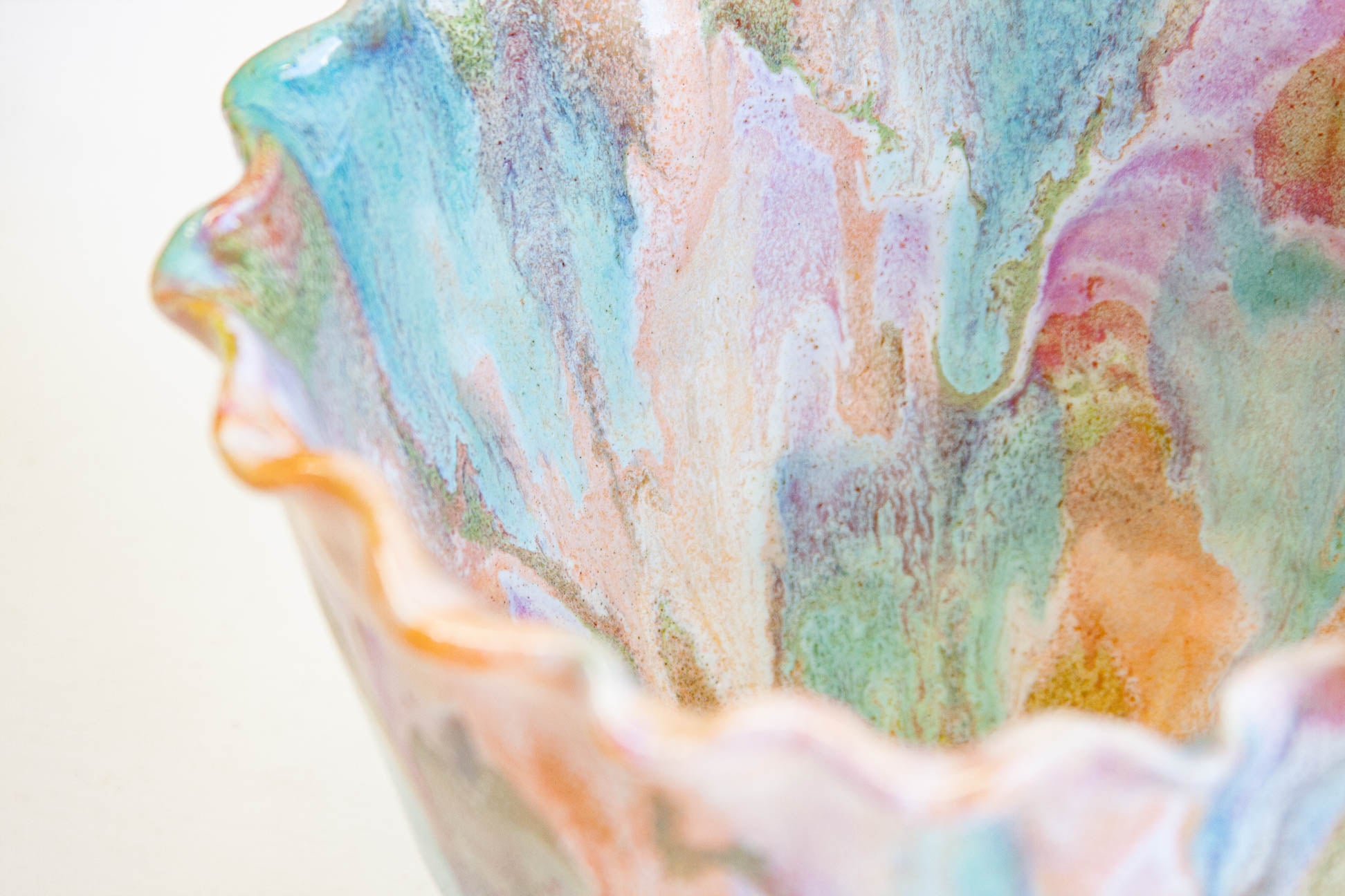 Close-up of the frilled rim of a handmade ceramic frill vessel, peach catching along the folded edge above green, blue and lilac.