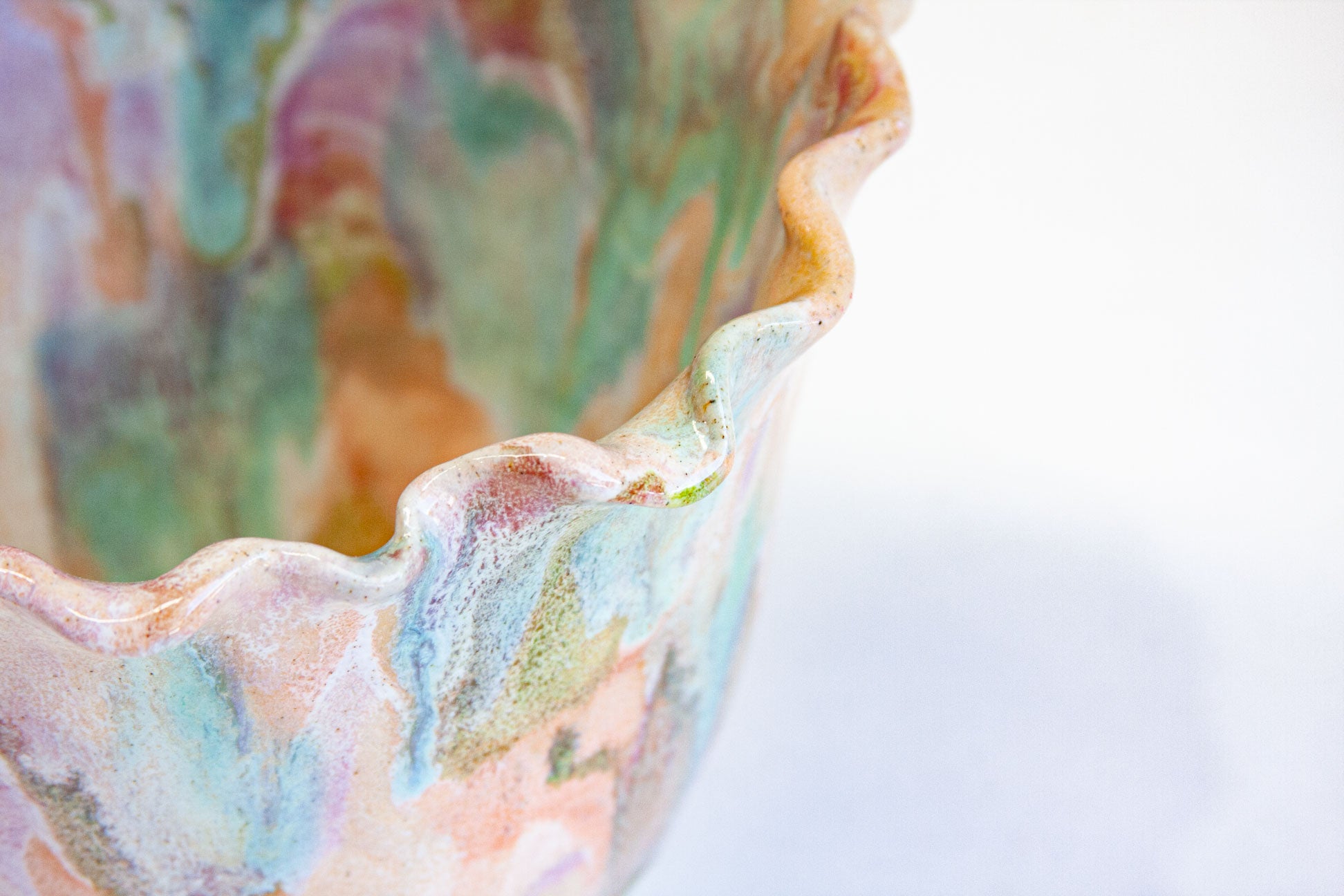 Close-up of the frilled rim of a handmade ceramic frill vessel, the folds glazed in peach, green and blue over soft lilac.