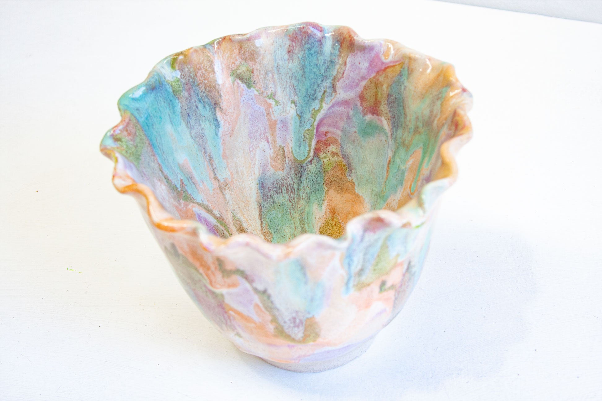A handmade ceramic frill vessel seen from above and to one side, the frilled rim curving around green, peach, blue and purple glaze.