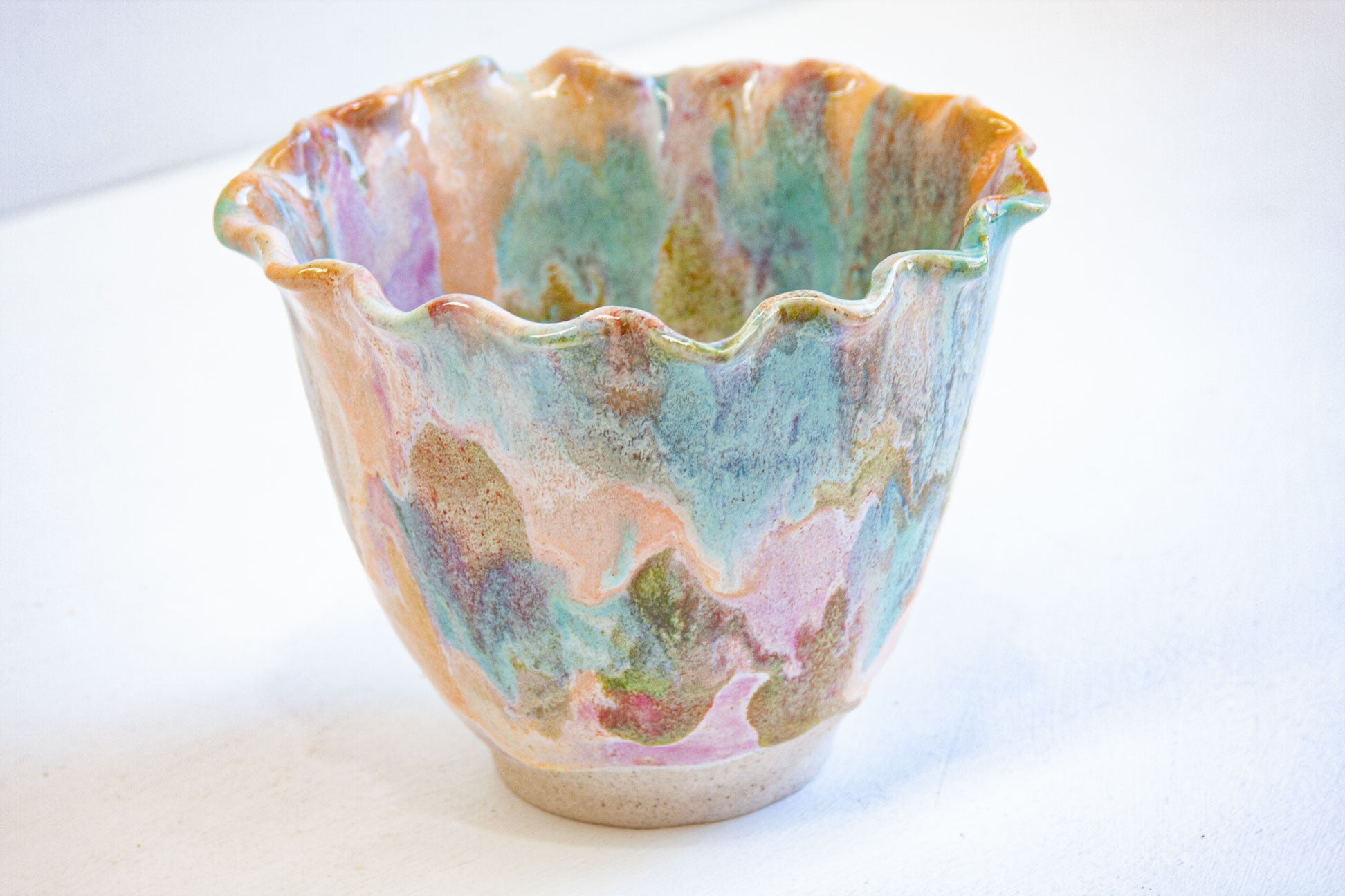 A handmade ceramic frill vessel seen at an angle, purple, green, peach and blue running down to a bare white clay foot.