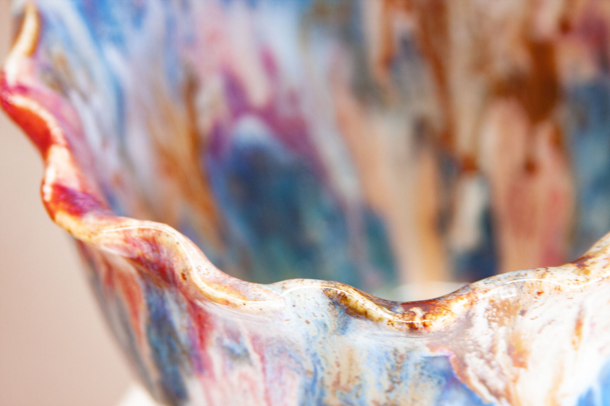 Close-up of the frilled rim of a handmade ceramic frill vessel, rust catching along the folded edge above pink, blue and white.
