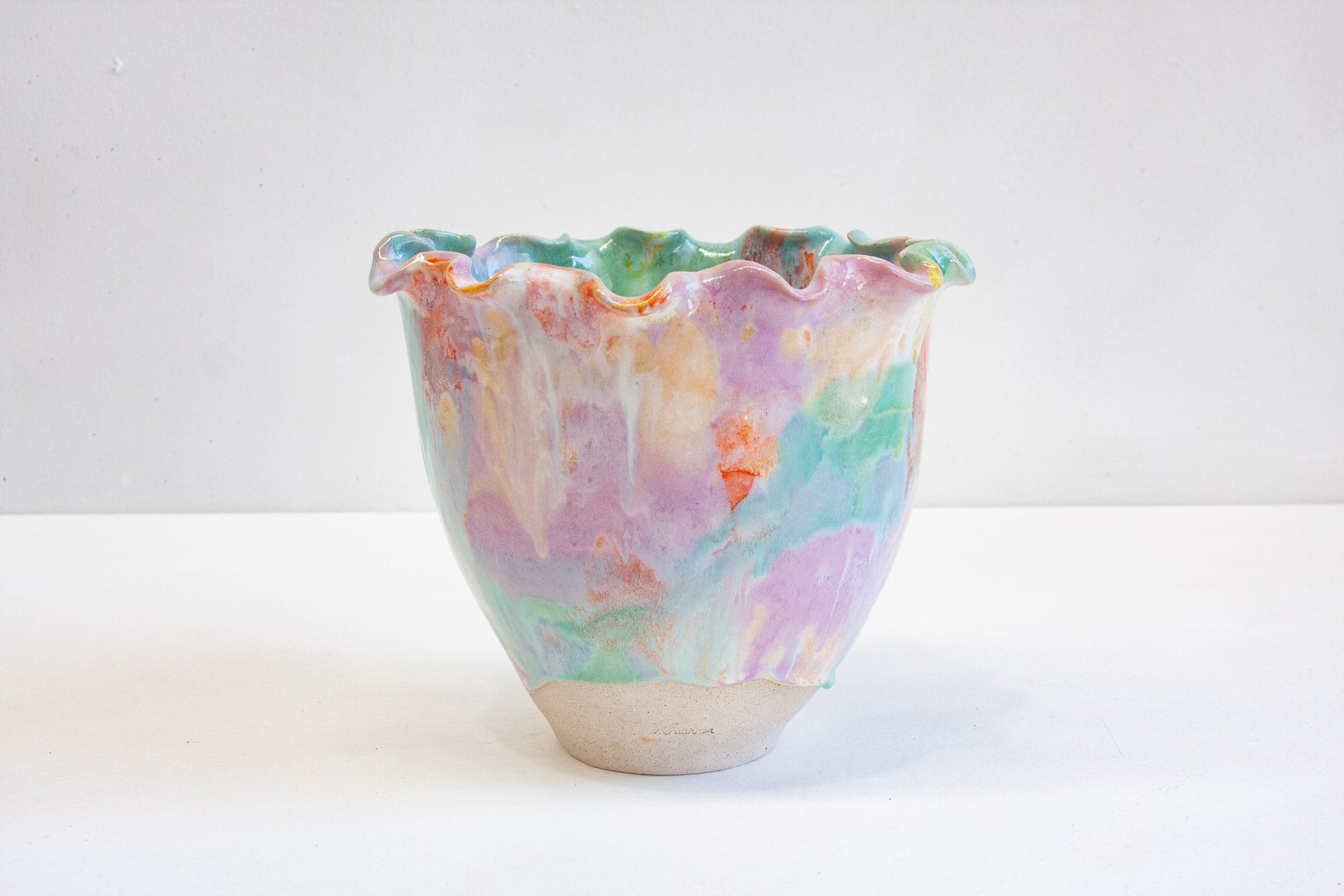 A handmade ceramic frill vessel seen from the front, glazed in purple, green, orange and yellow above a bare white clay foot.