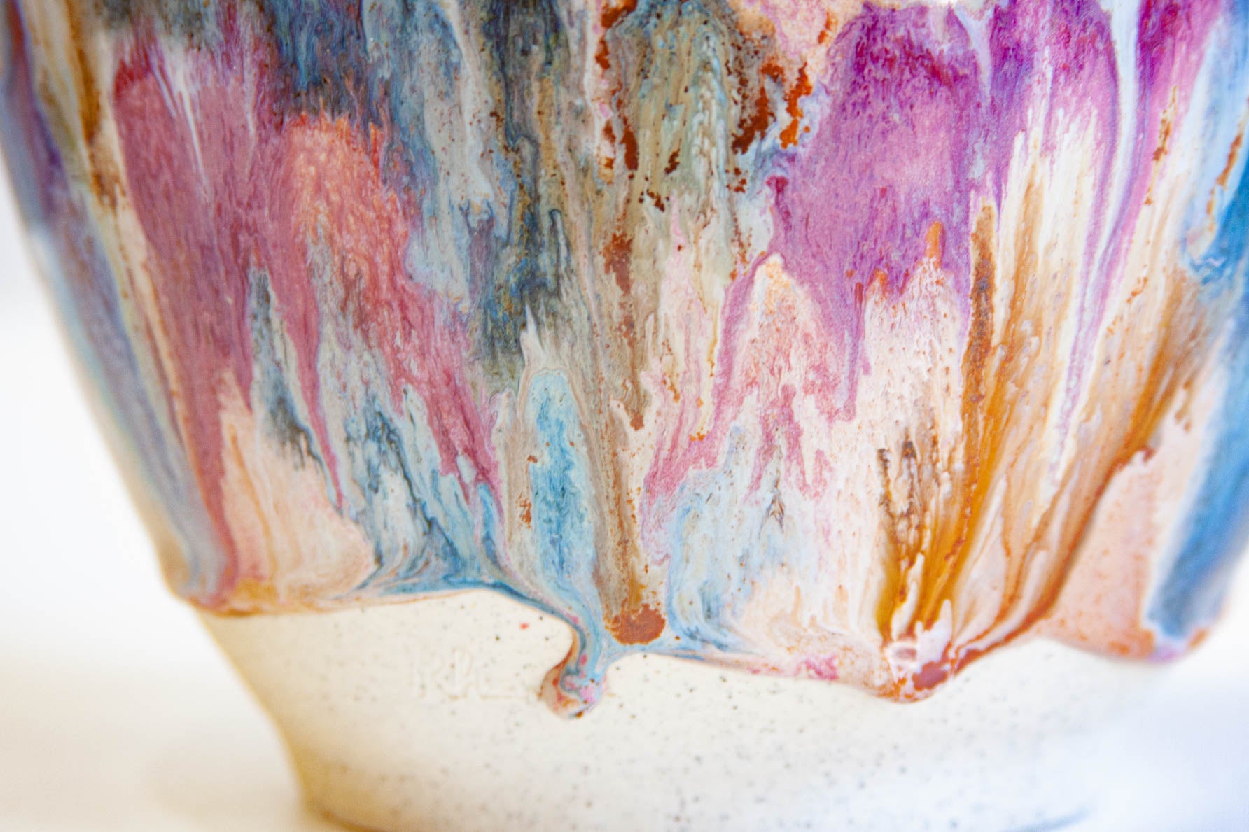 Close-up of the lower half of a handmade ceramic frill vessel, pink, blue and rust running into drips above the bare white clay foot.