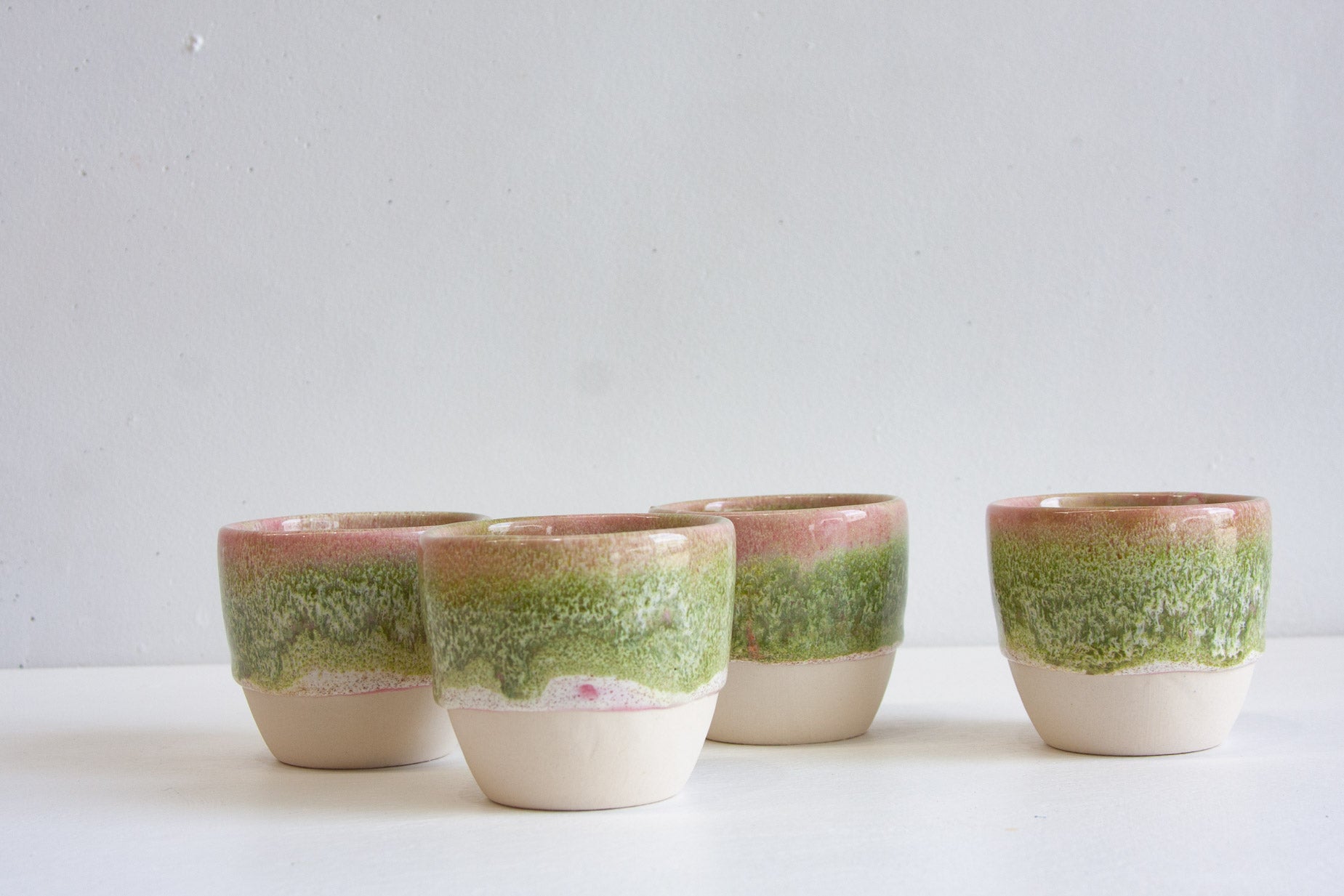 Four handmade ceramic cups grouped together, each with a pink rim running into green above a bare white clay base.