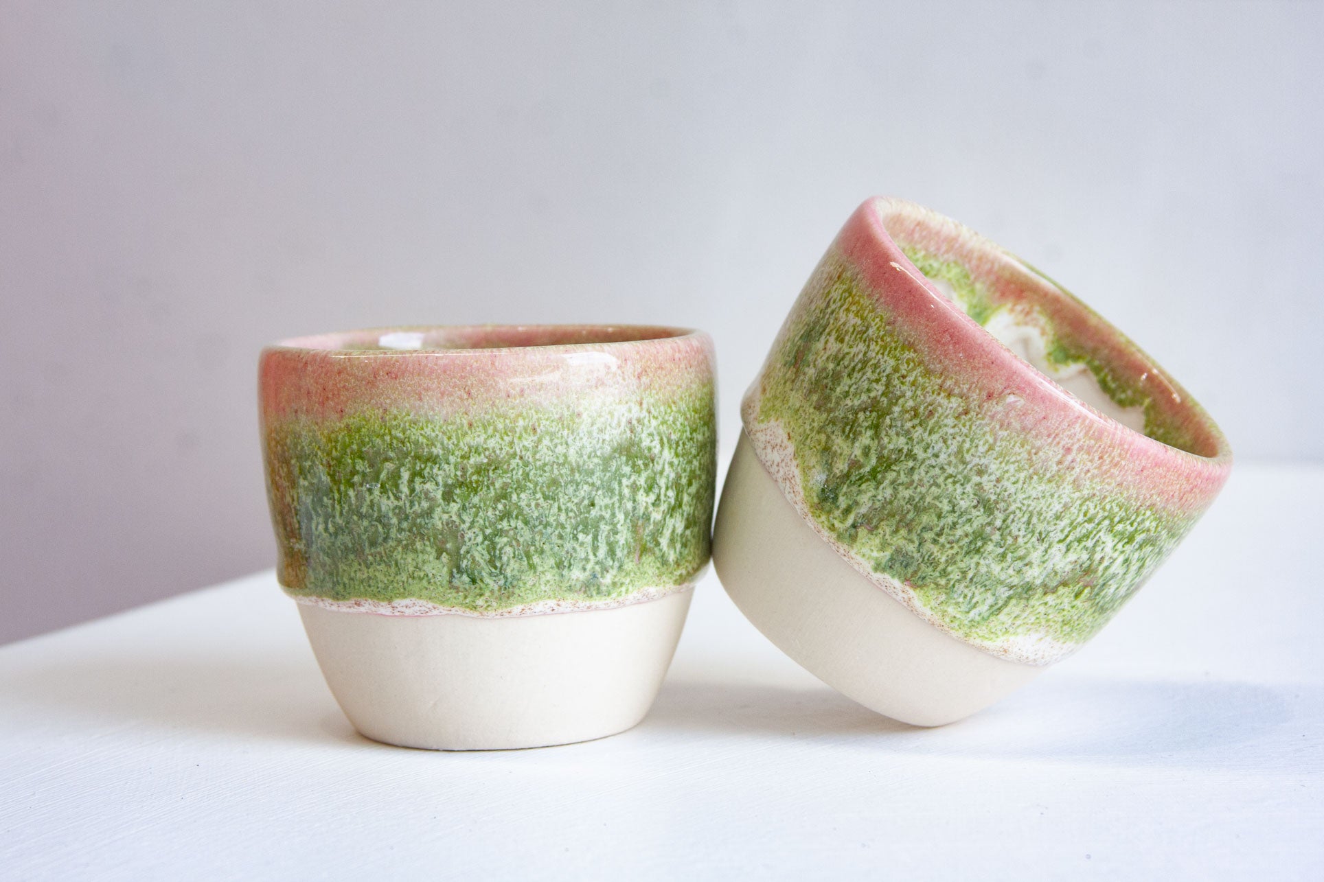 Two handmade ceramic cups, one tipped on its side, pink rims fading into mossy green over bare white clay.