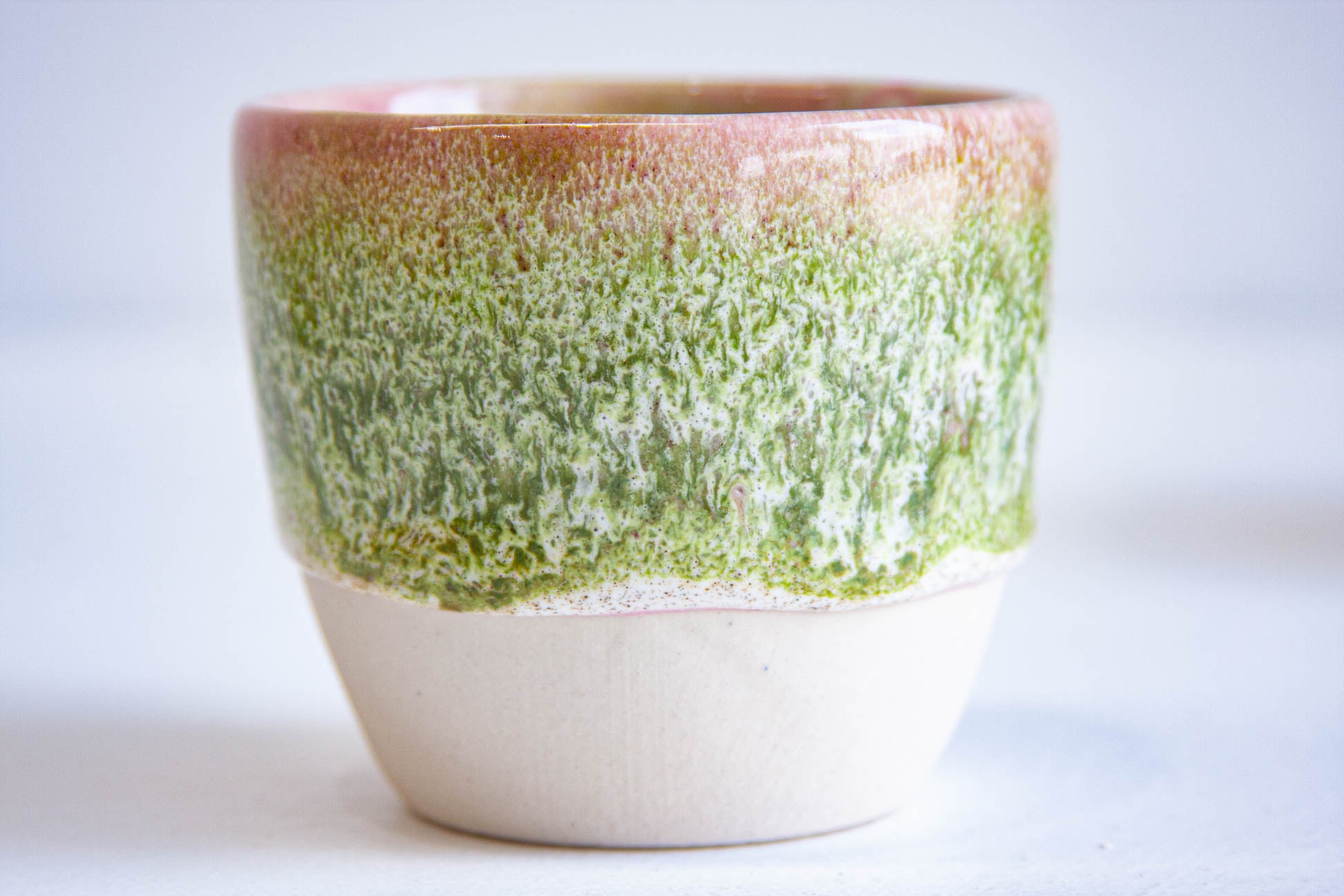 Close-up of a single handmade ceramic cup, pink at the rim breaking into dense mossy green above the bare white clay.