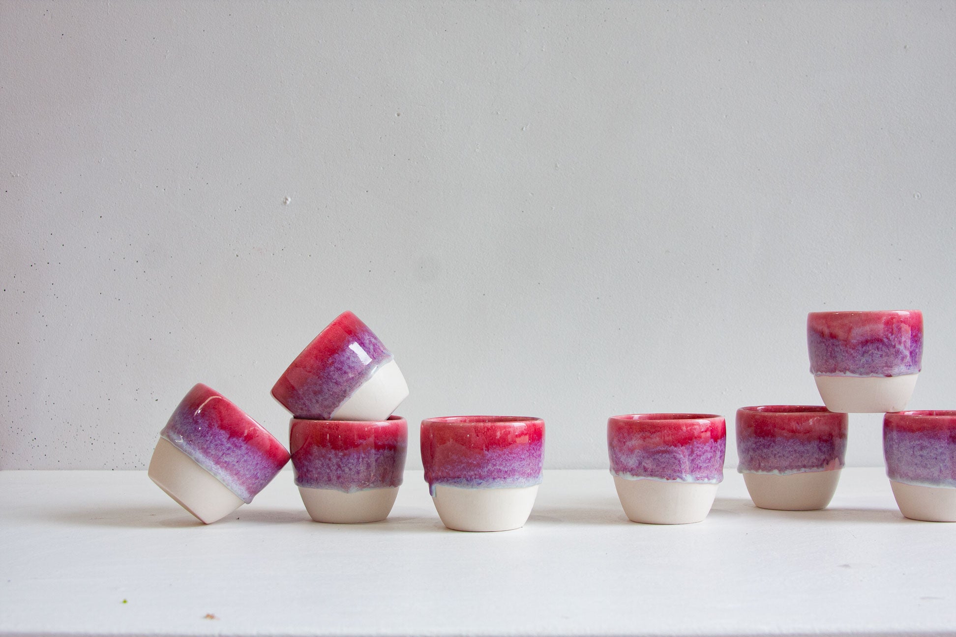 Eight handmade ceramic cups arranged in a group, dip-glazed in deep pink fading through violet speckle to a bare white clay base.