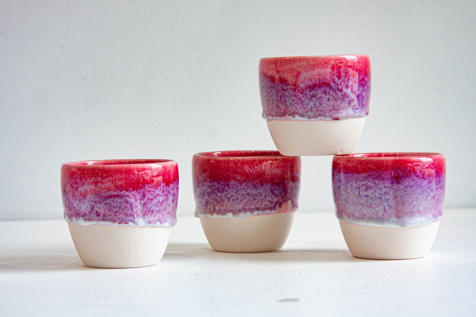 Four handmade ceramic cups stacked in a pyramid, each dip-glazed in deep pink fading through violet to bare white clay.
