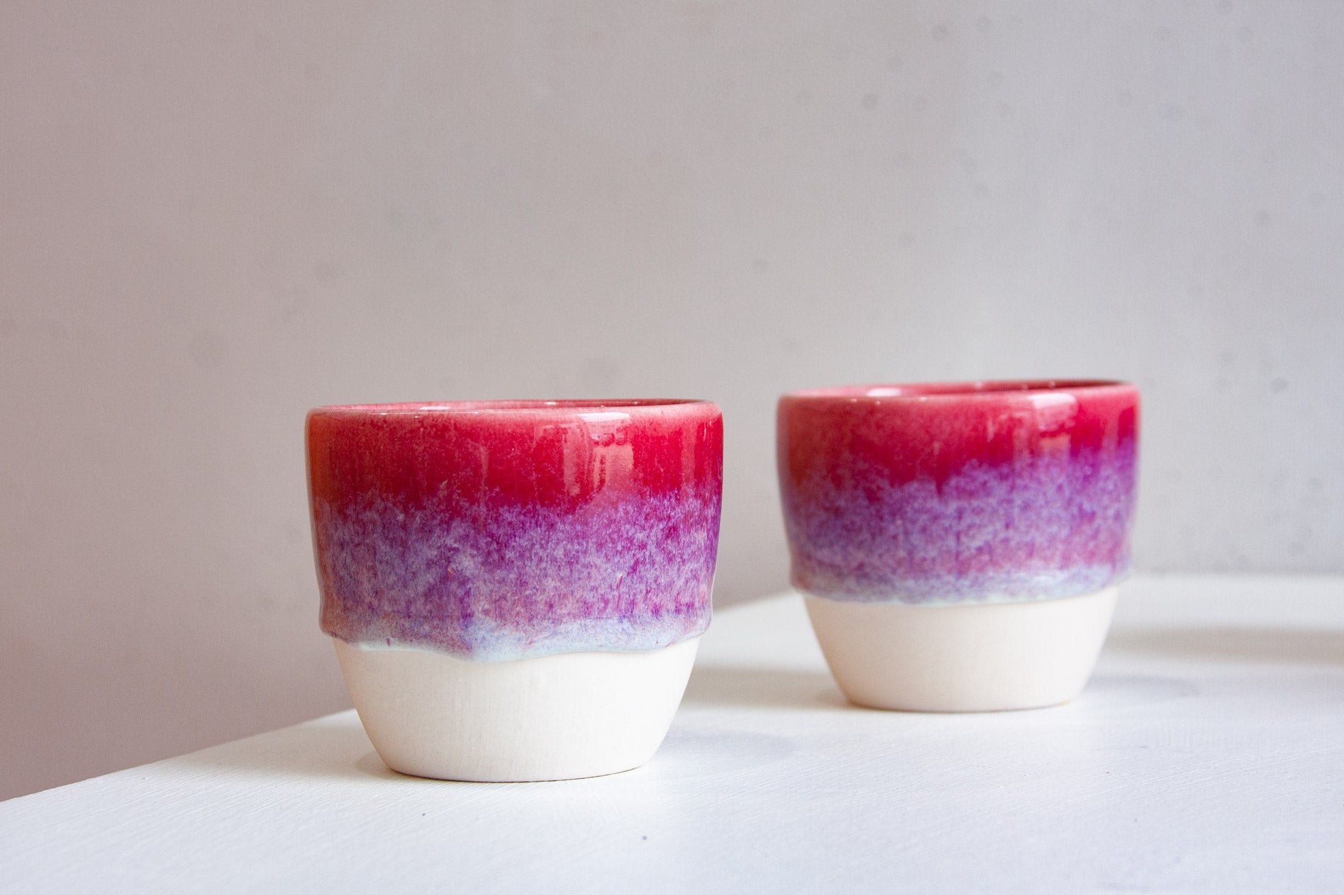 Two handmade ceramic cups side by side, the deep pink glaze breaking into violet speckle before it stops above the bare white clay.