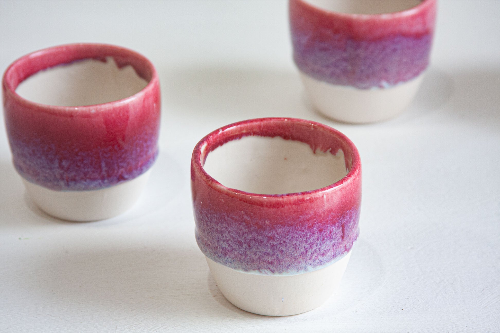 Three handmade ceramic cups seen from above at an angle, the deep pink rim dropping into violet speckle above the bare white clay.