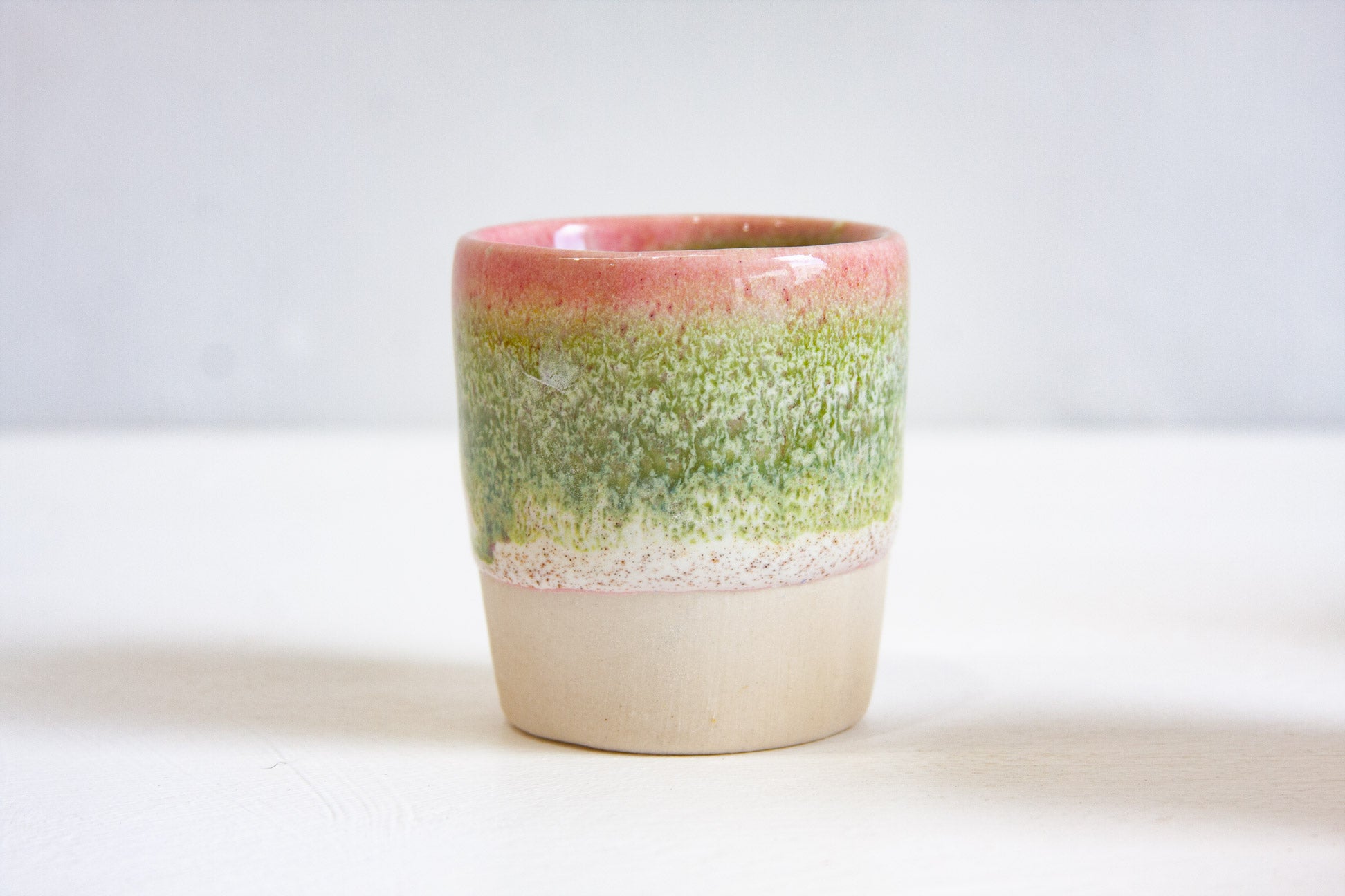 A single handmade ceramic espresso cup front on, a pink rim fading into speckled green above the bare white clay base.