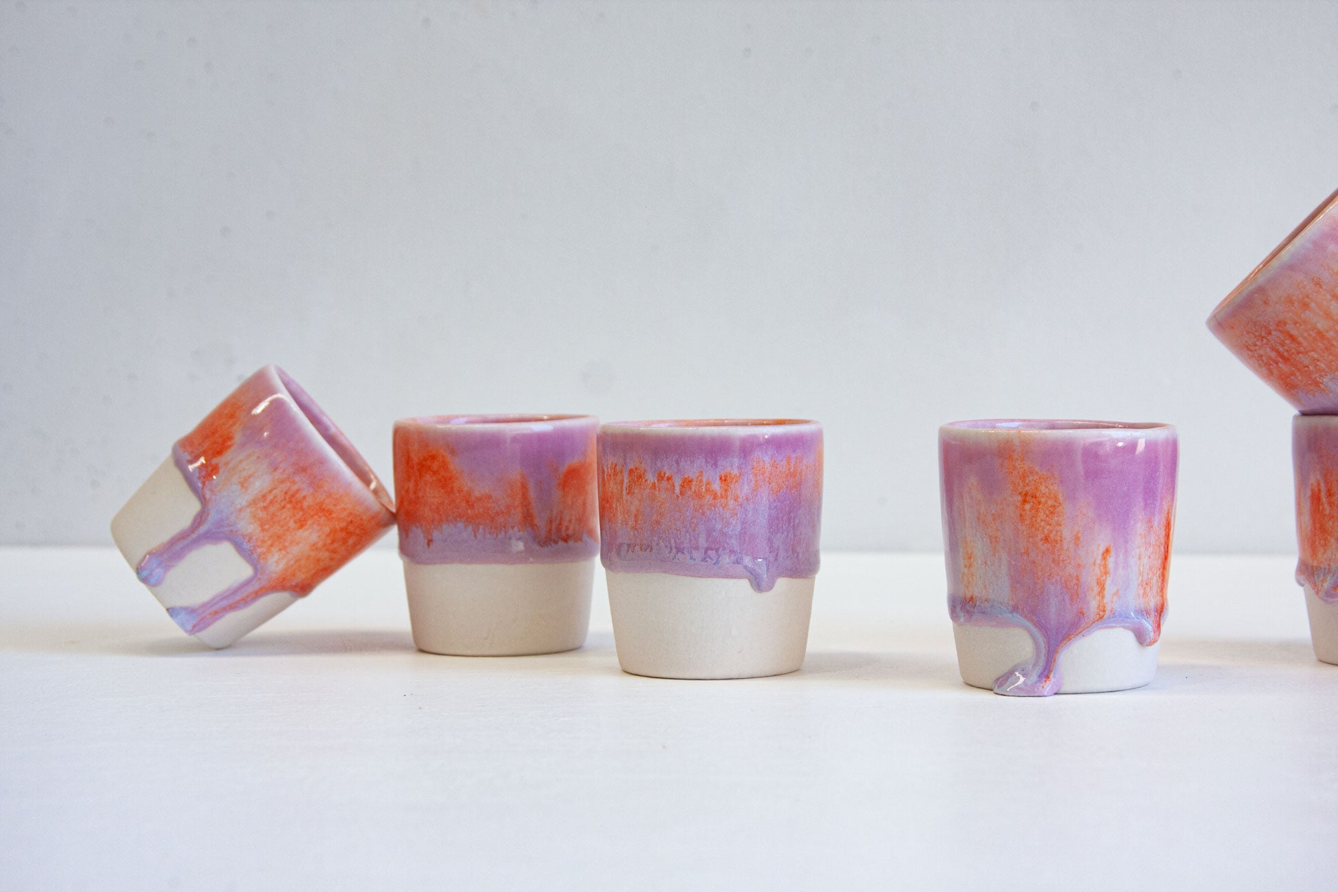 Six handmade ceramic espresso cups grouped together, one tipped on its side, each glazed in purple and orange over bare white clay.