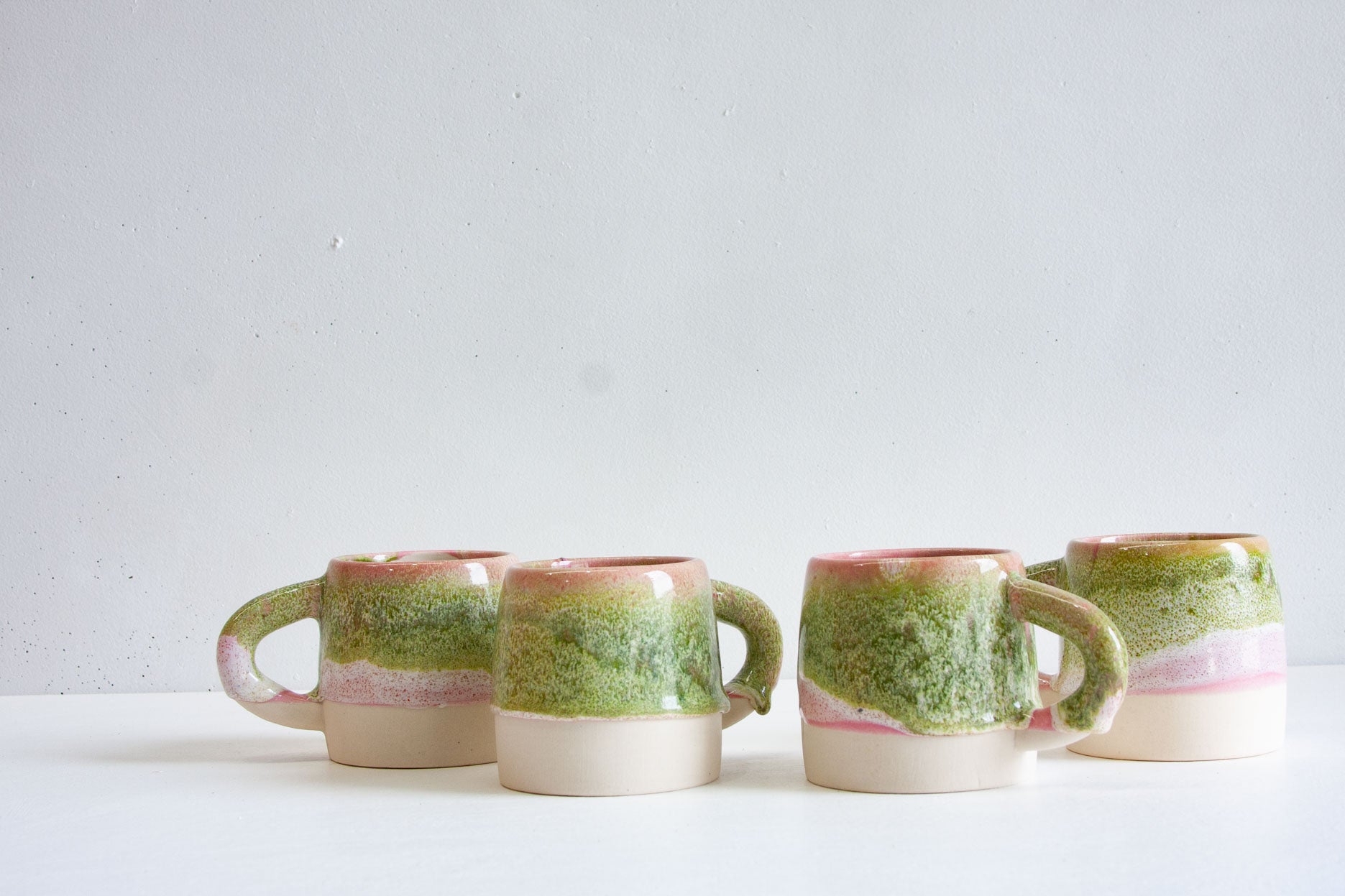 Four handmade ceramic large mugs in a pink and green glaze lined up in a row on a white table.