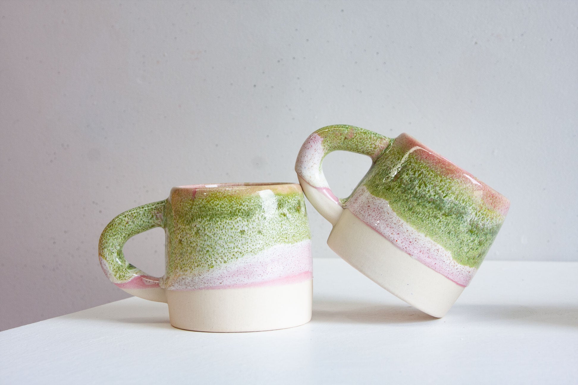 Two handmade ceramic large mugs in a pink and green glaze, one standing upright and one tipped on its side.