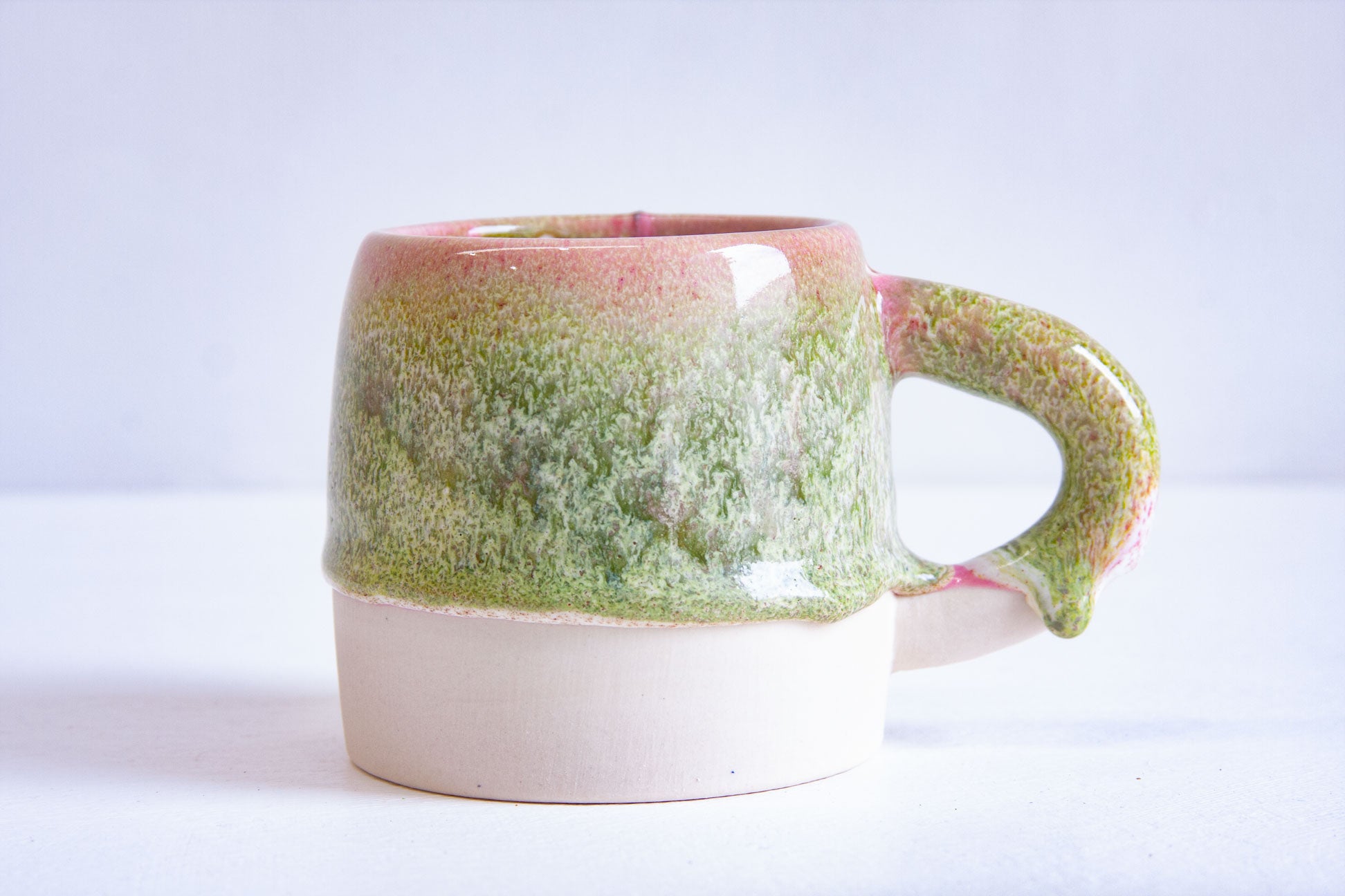 A handmade ceramic large mug in a pink and green glaze over bare white clay, handle to the right.