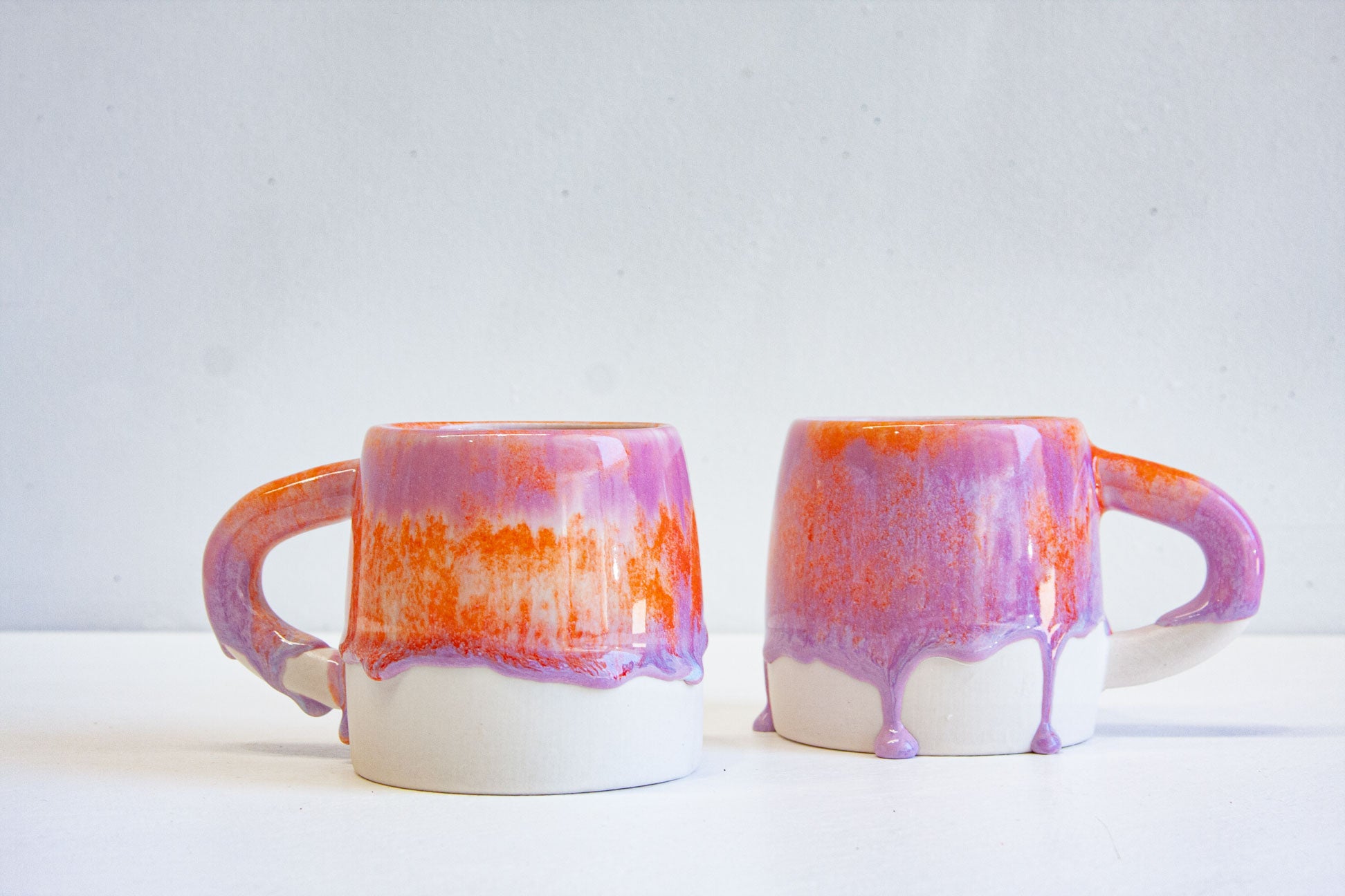 Two handmade ceramic large mugs side by side, handles out, orange over purple with the glaze running into drips above the bare clay base.