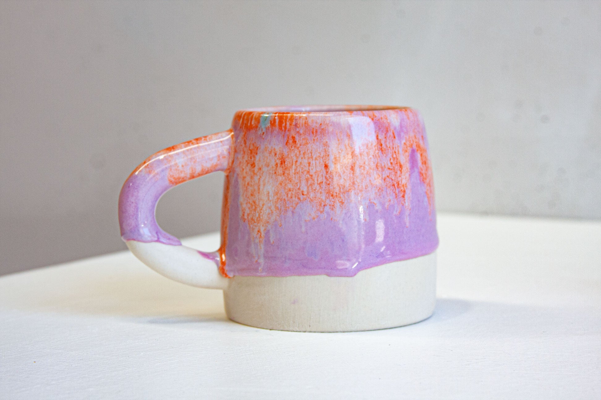 A single handmade ceramic large mug seen side on, orange breaking across a purple glaze that runs in drips down to the bare clay base.