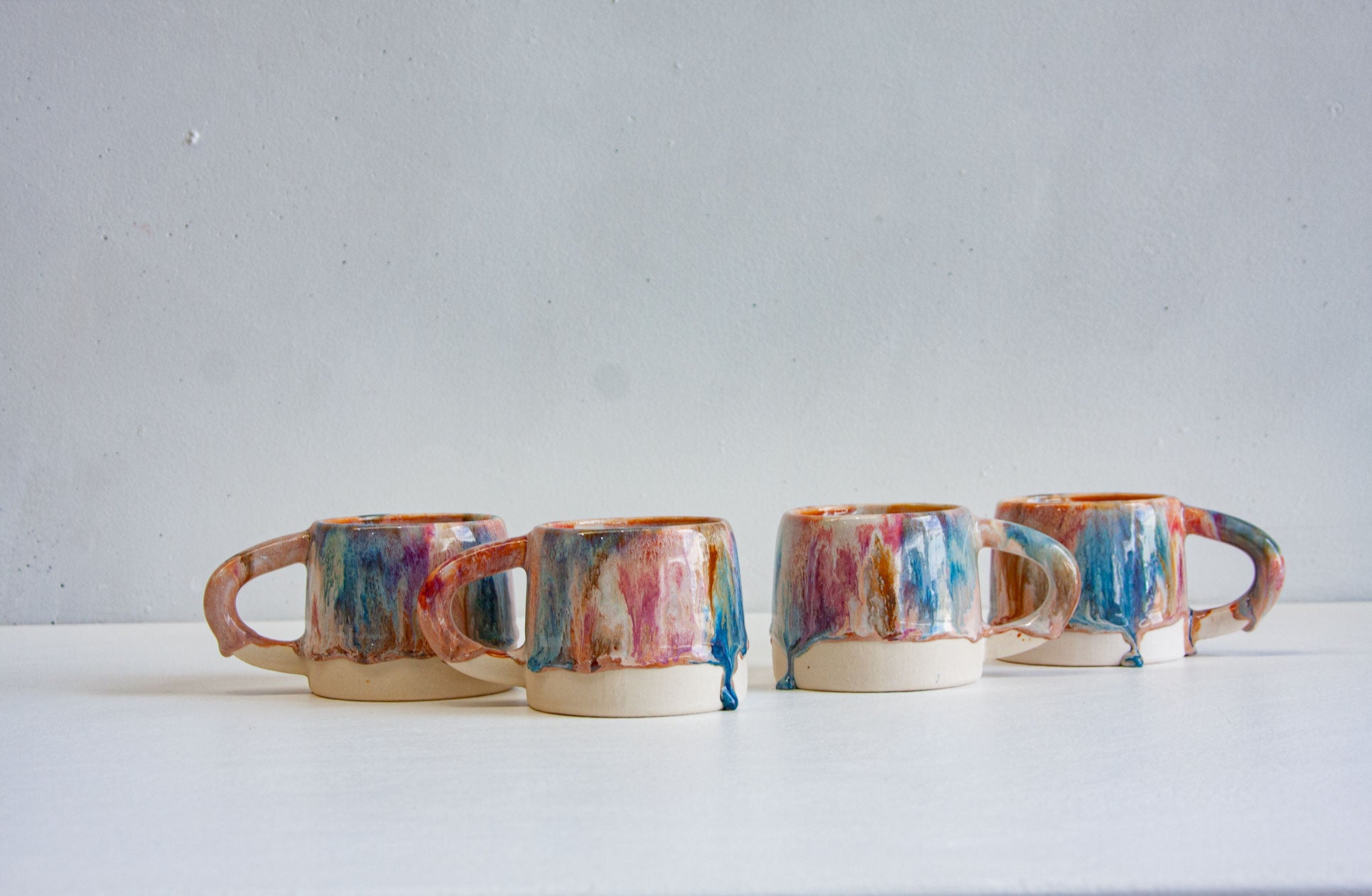 Four handmade ceramic mugs in a row, handles out, the brown, pink and blue glaze running in vertical streaks to a bare white clay base.