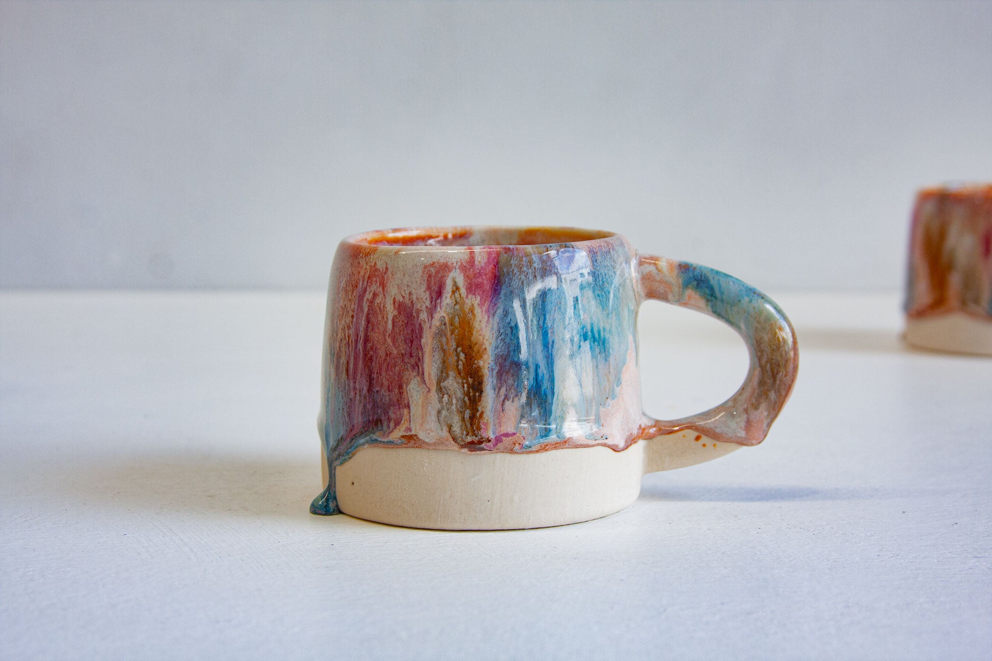 A single handmade ceramic mug seen side on, layers of brown, pink and blue falling to an uneven line above the bare white clay.