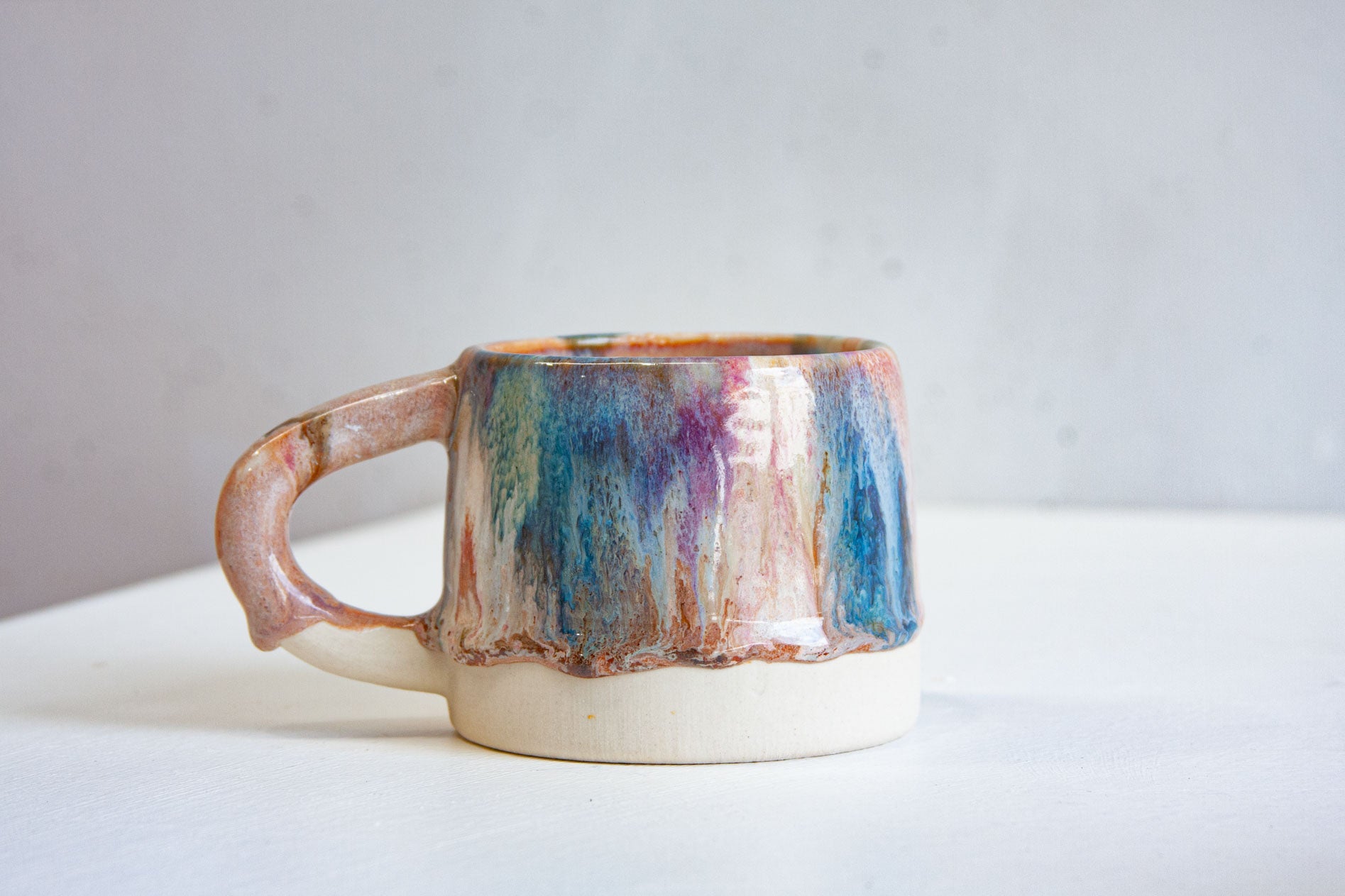 A handmade ceramic mug with the handle to the left, brown, pink and blue layered down the body over bare white clay.