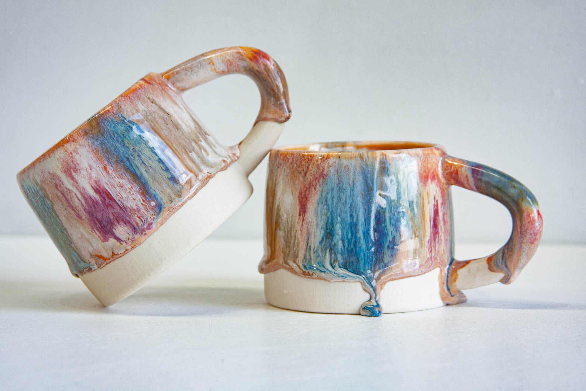 Two handmade ceramic mugs, one tipped on its side, both glazed in layered brown, pink and blue above bare white clay bases.