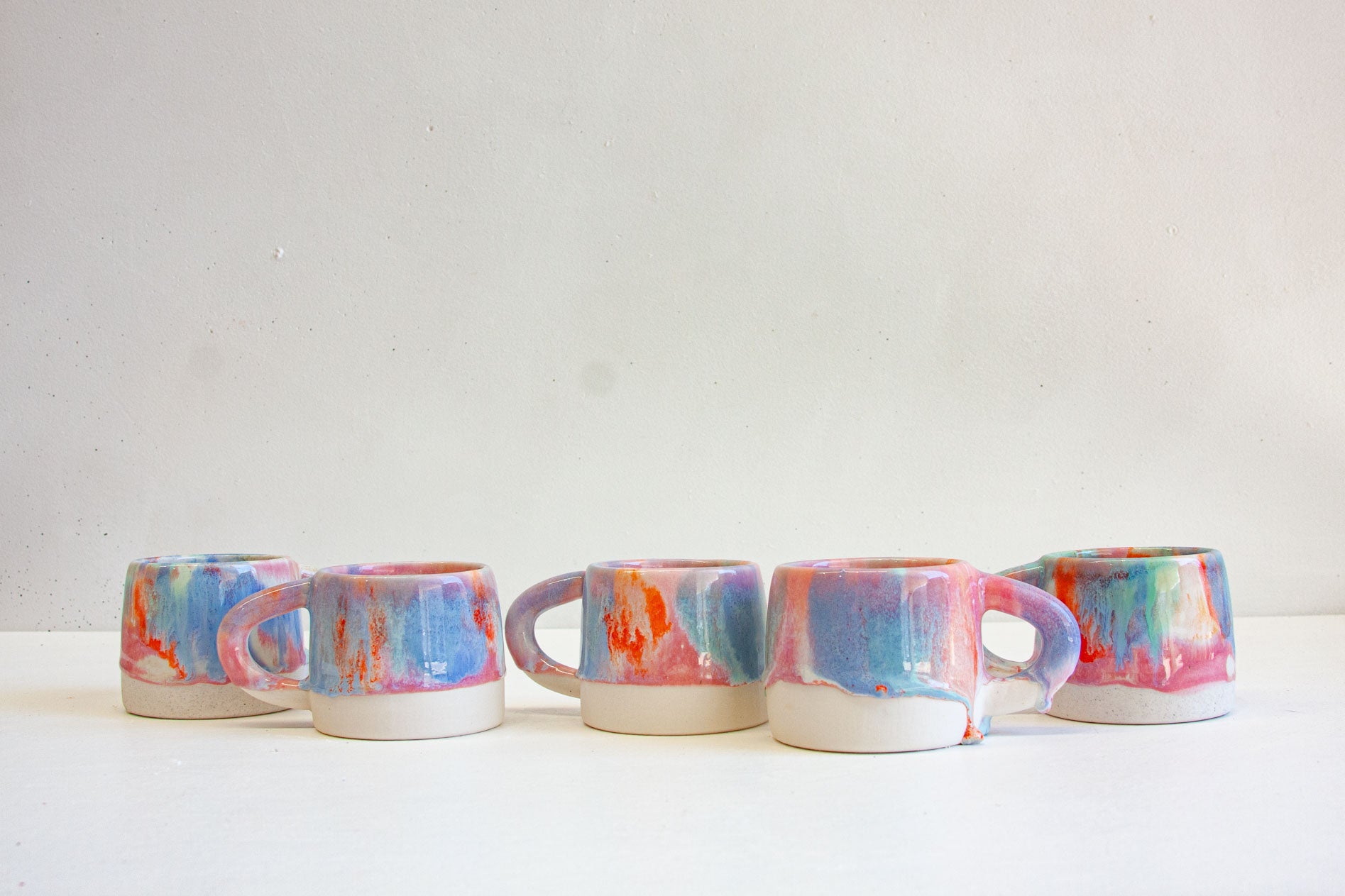 Five handmade ceramic mugs in a row, each glazed in pink, blue, orange and green over a bare white clay base.