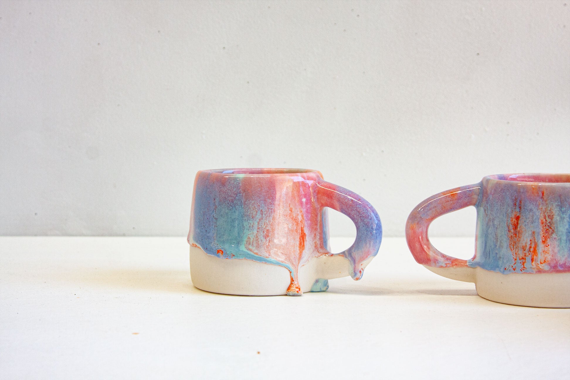Two handmade ceramic mugs side on, the pink, blue and orange glaze ending in a line of drips above the bare white clay base.