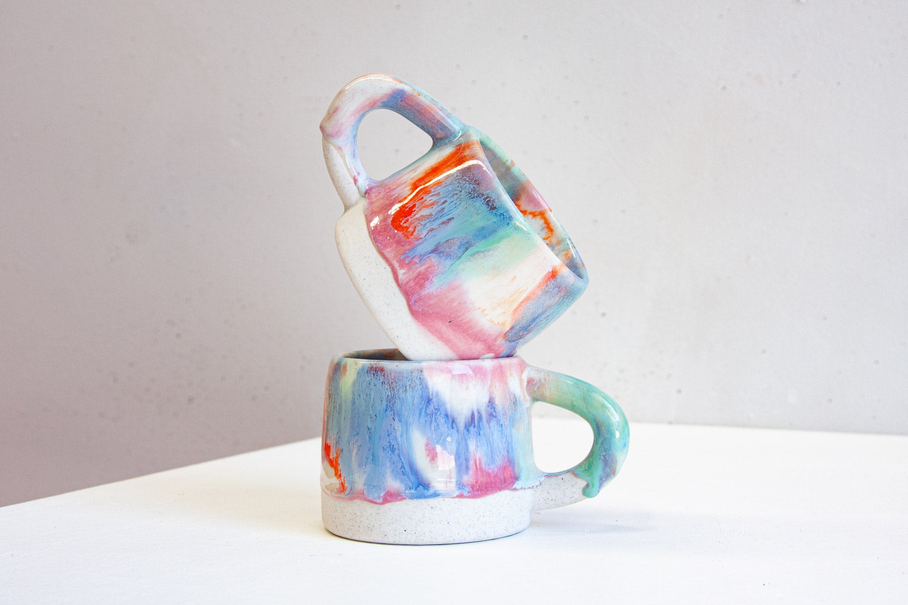Two handmade ceramic mugs stacked, the upper one tipped to show pink, blue, orange and green glaze over bare white clay.