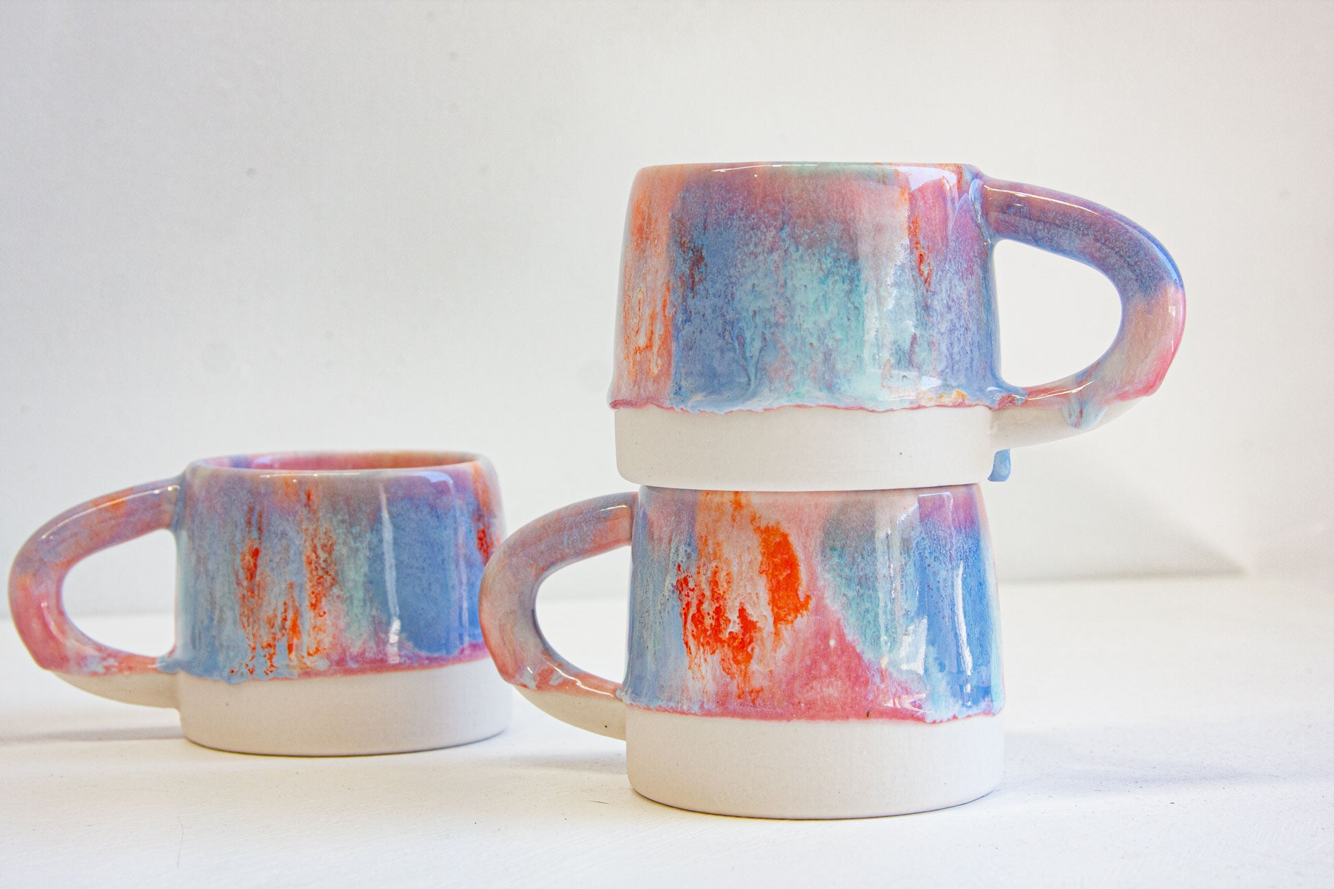 Three handmade ceramic mugs, two stacked and one beside them, glazed in pink, blue and orange over bare white clay.