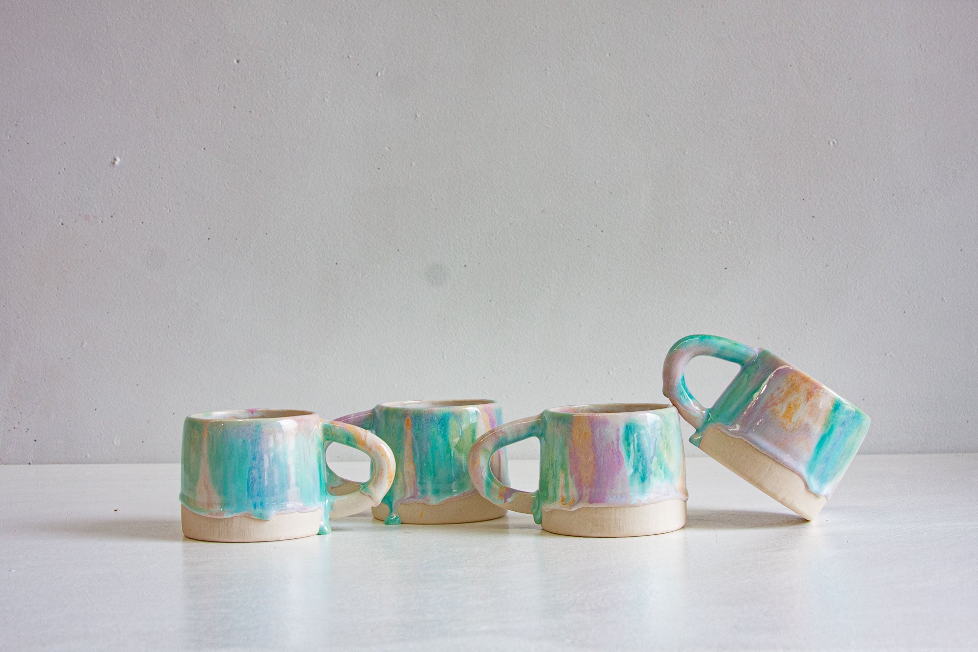 Four handmade ceramic mugs in Sorbet, mint green, purple and peach glaze running into drips over bare white clay, one tipped on its side.