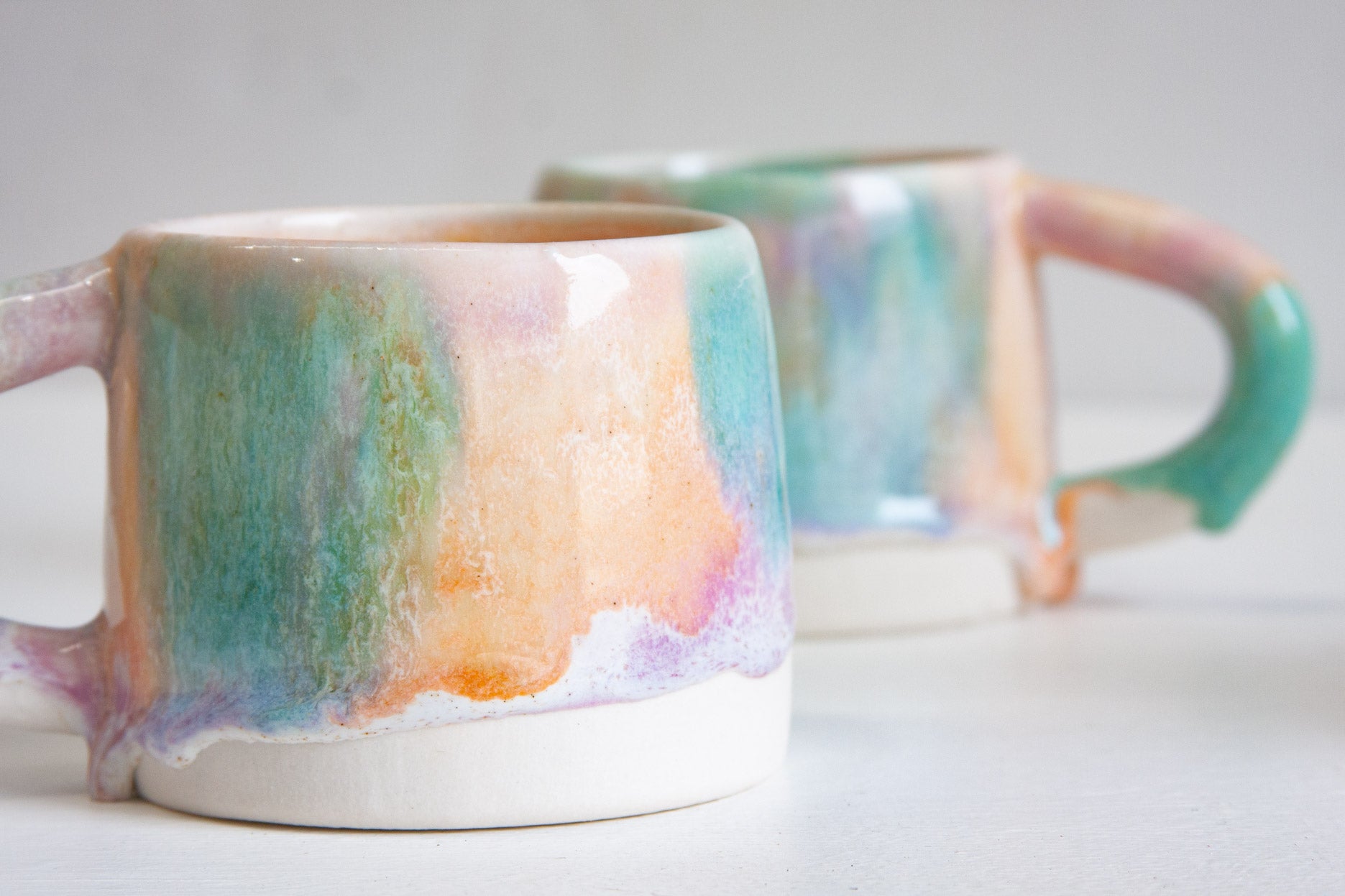 Two handmade ceramic mugs, the front one angled to show the pastel purple, green and orange glaze pooling above the bare white clay base.