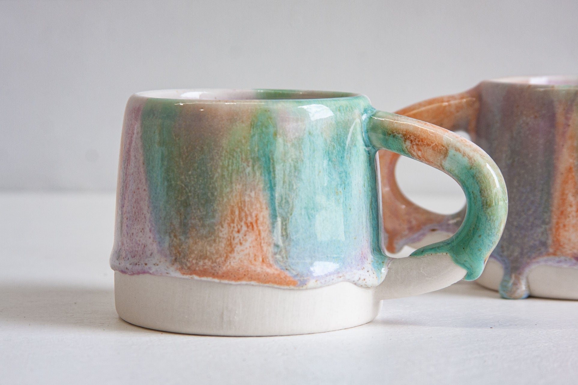 Close-up of a handmade ceramic mug with a green glazed handle, the orange and purple glaze trailing down over the bare white clay base.