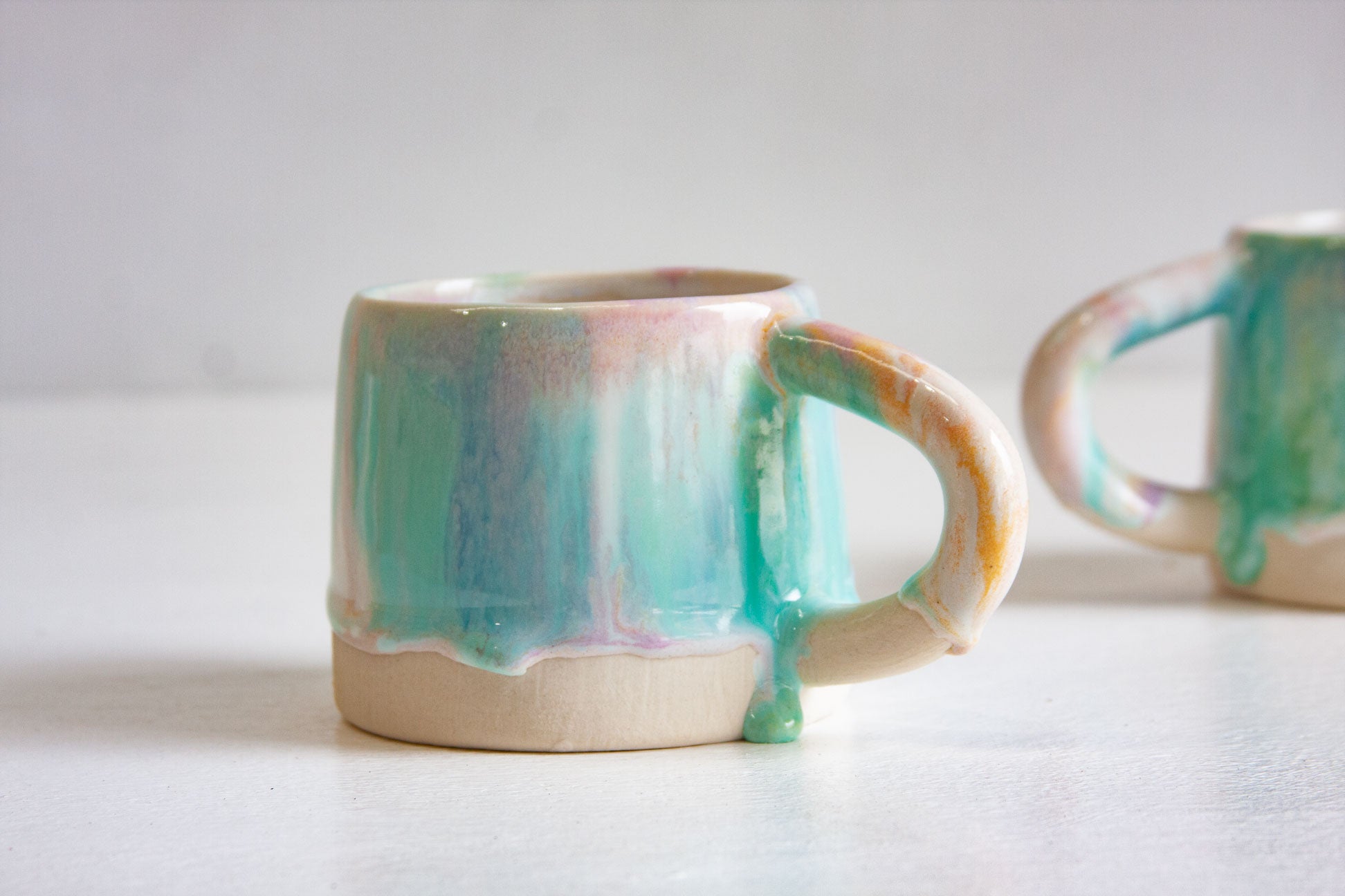 A handmade ceramic mug in Sorbet, handle to the right, mint green and purple glaze ending in drips above the bare white clay base.