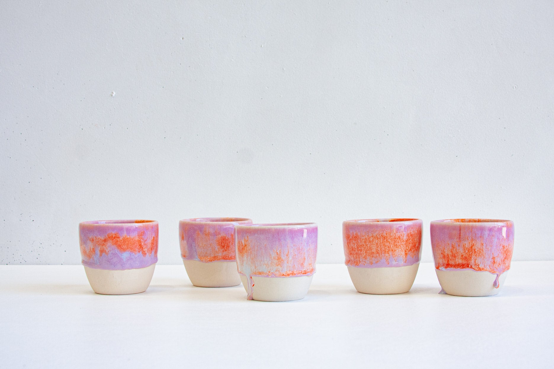 Five handmade ceramic cups in a row, dip-glazed in purple with a band of vivid orange, stopping above the bare white clay base.