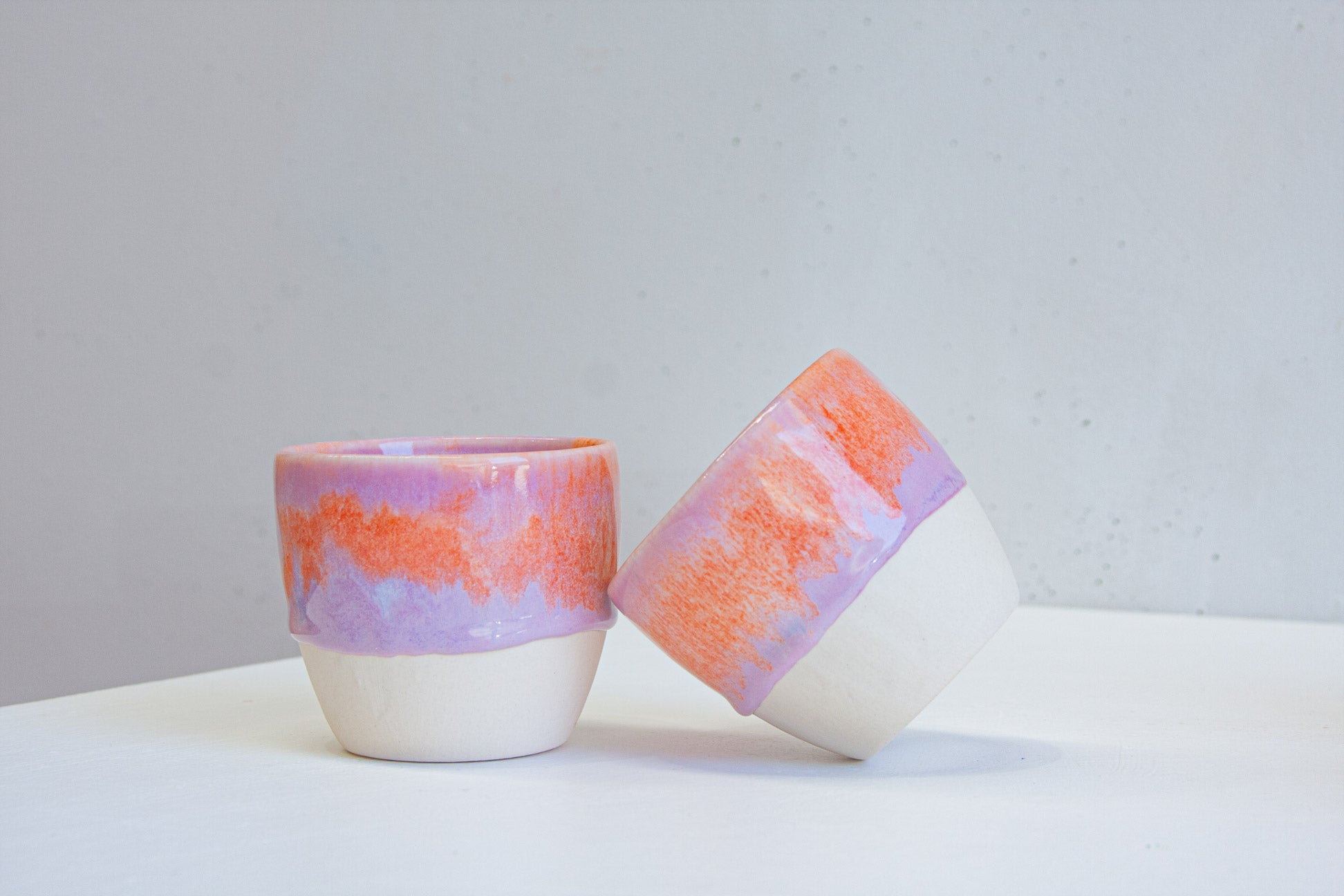Two handmade ceramic cups, one tipped on its side, the purple and orange glaze ending in an uneven line above the bare white clay.