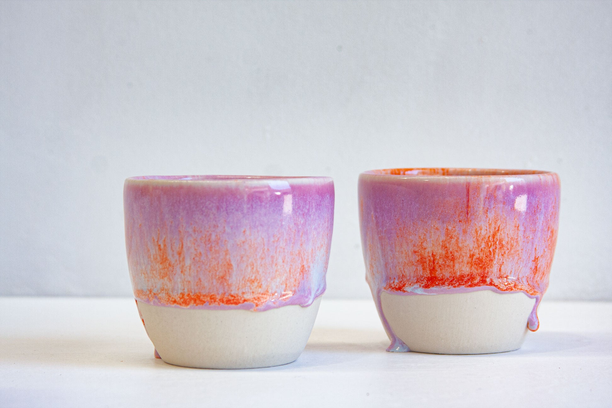 Two handmade ceramic cups side by side, orange breaking through the purple glaze and running into small drips above the bare clay.