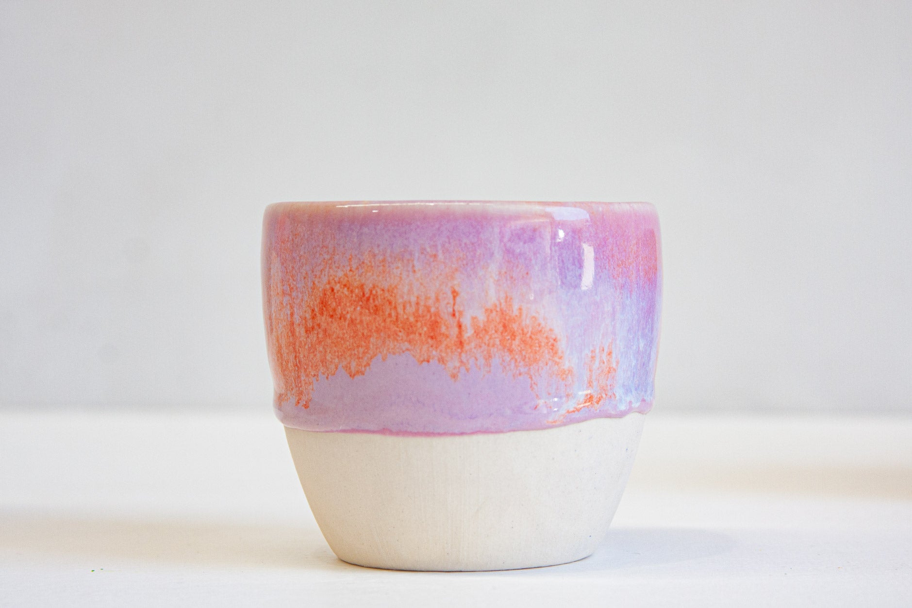 A single handmade ceramic cup front on, a band of vivid orange across the purple glaze above the bare white clay base.
