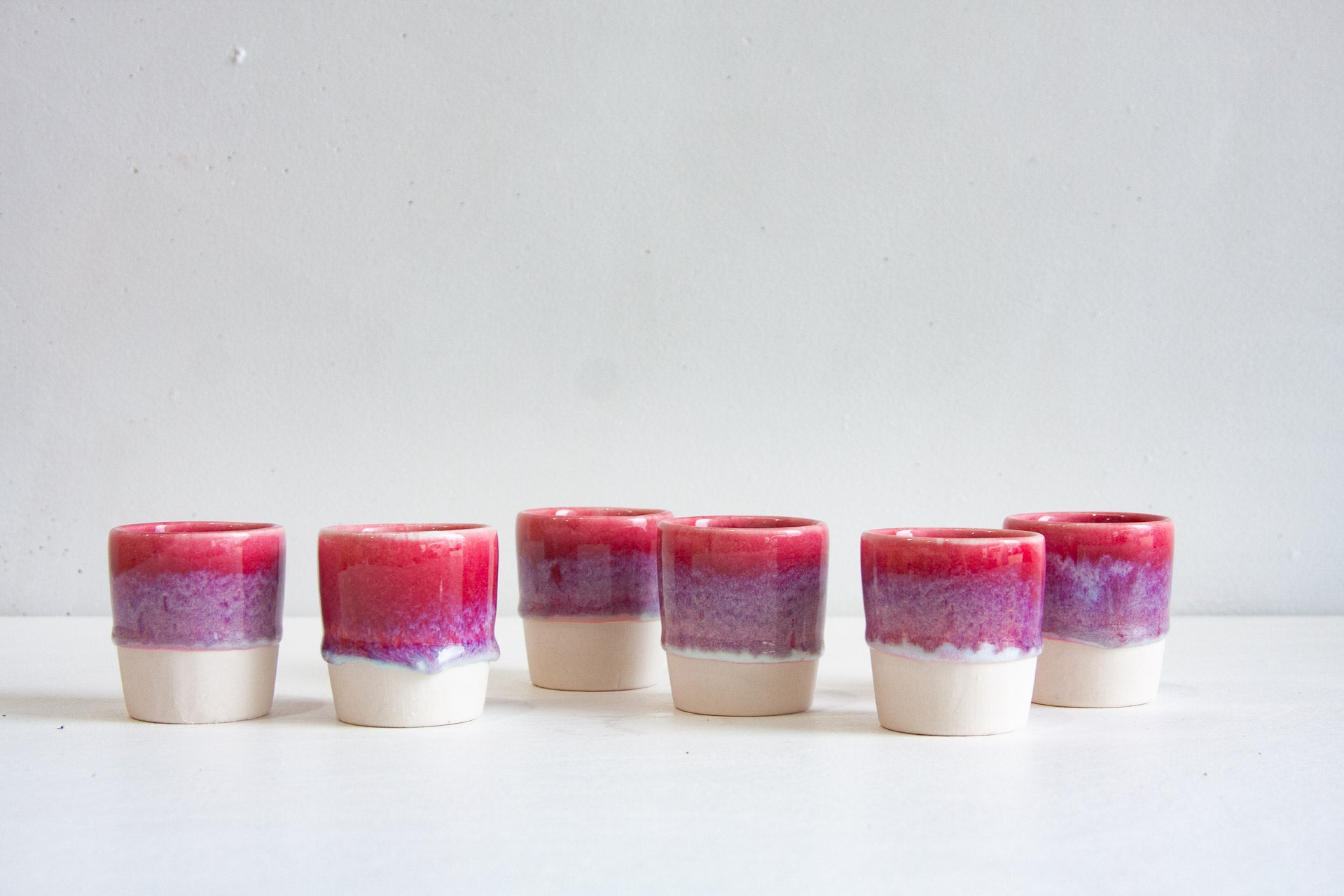 Six handmade ceramic espresso cups arranged in a group, each dip-glazed in deep pink fading into lilac above a bare white clay base.