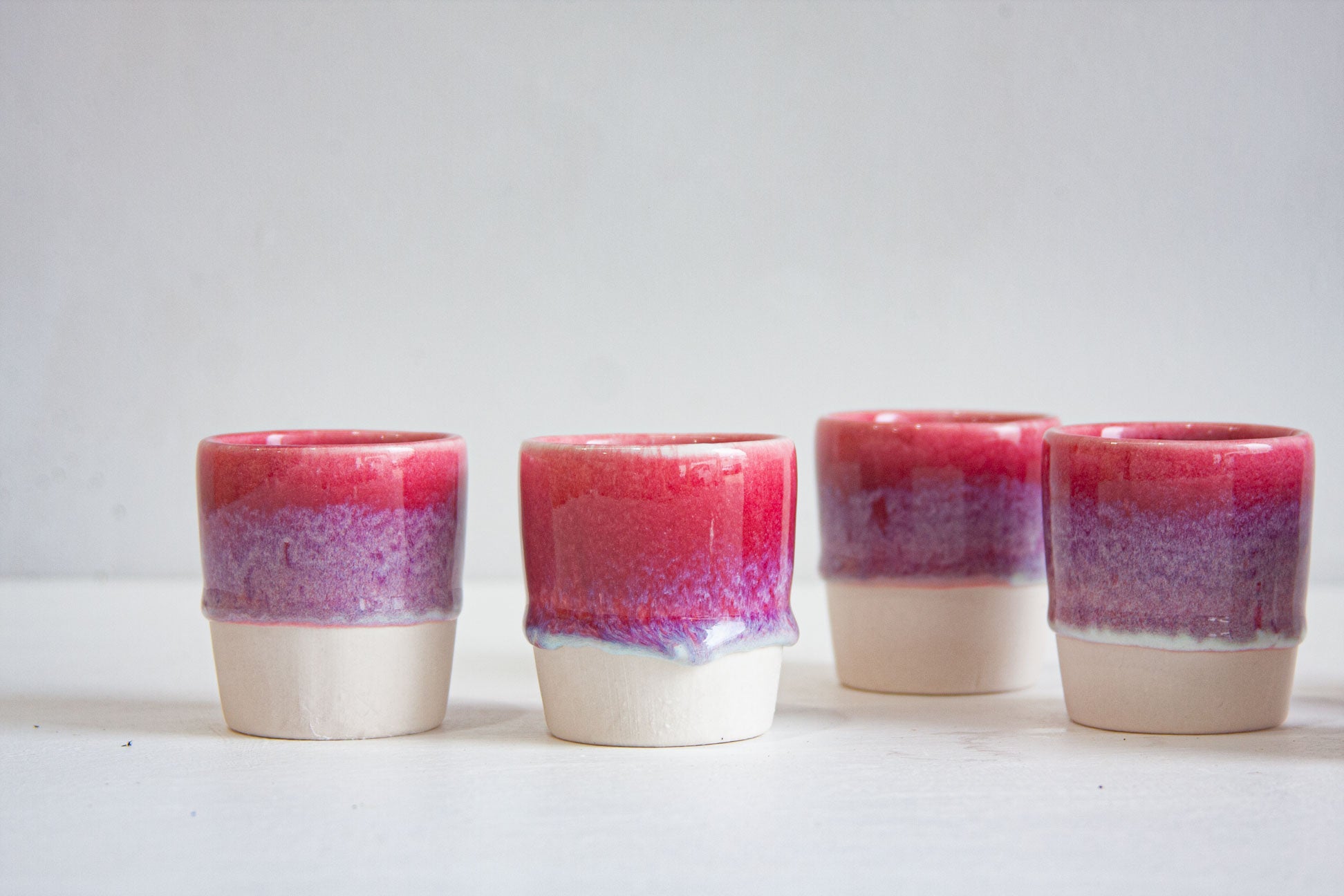 Four handmade ceramic espresso cups in a row, dip-glazed in deep pink fading through lilac speckle to a bare white clay base.