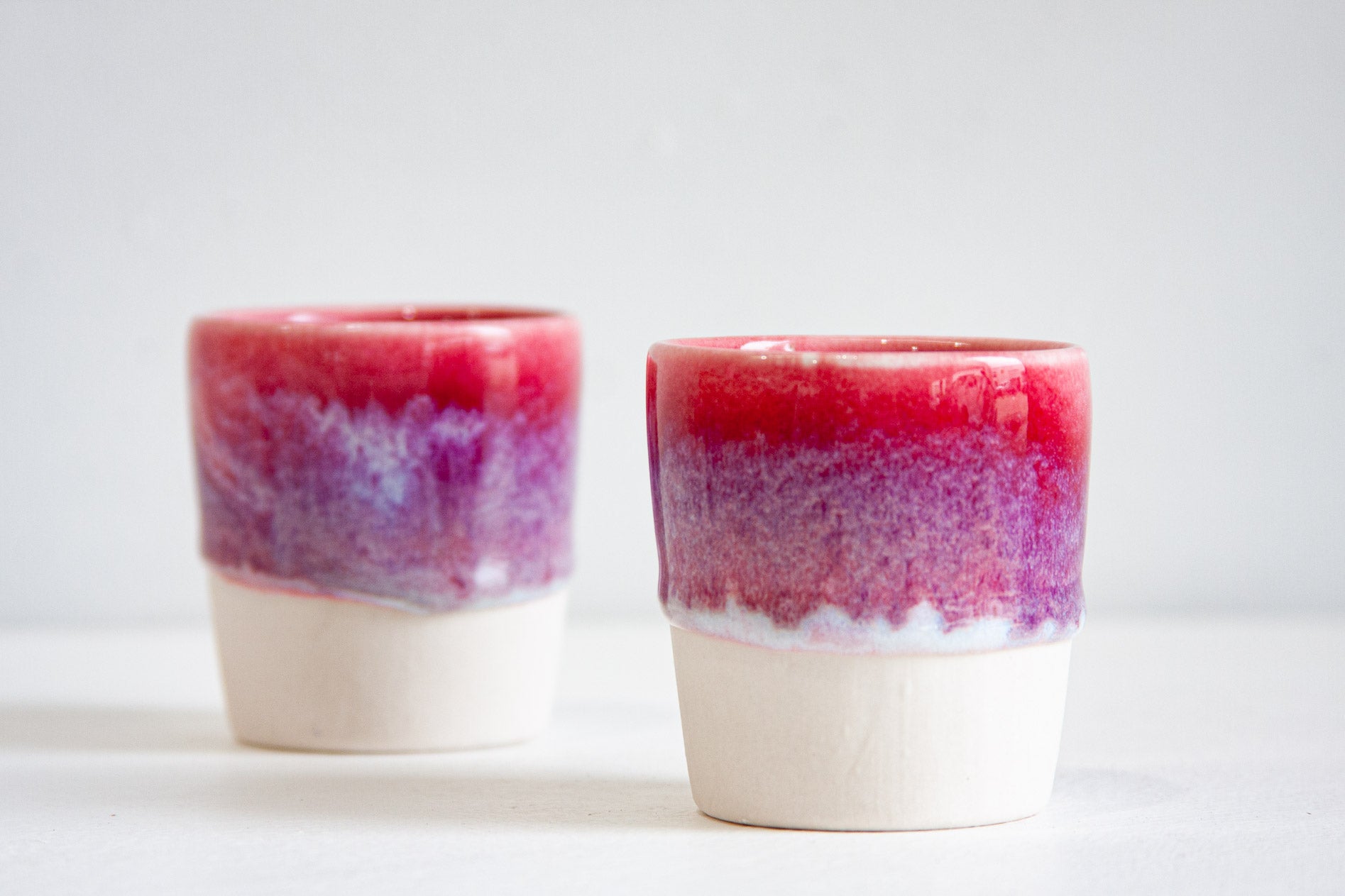 Two handmade ceramic espresso cups close up, the deep pink glaze breaking into lilac speckle before it stops above the bare white clay.