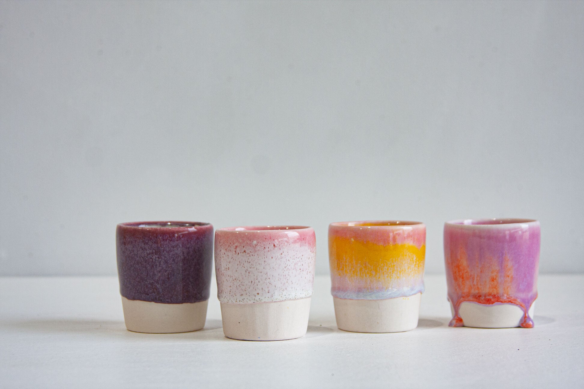Four handmade ceramic espresso cups in a row: a deep plum, a speckled white with a pink rim, a golden yellow with a lilac band, and a pink and lilac with orange drips.