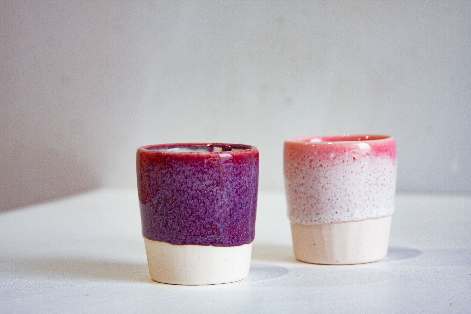 Two handmade ceramic espresso cups, one a deep speckled plum, the other a soft white flecked with pink beneath a bright pink rim, both on bare clay bases.