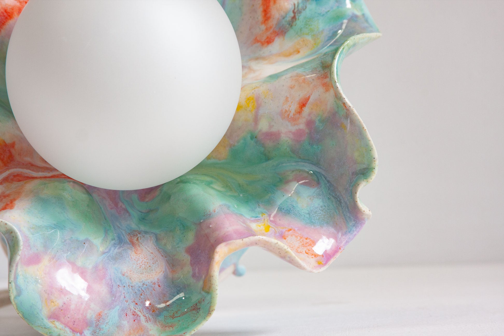 Close-up of one ruffled fold of a handmade ceramic Oyster table lamp beside the white globe, glazed in green, pink and orange.