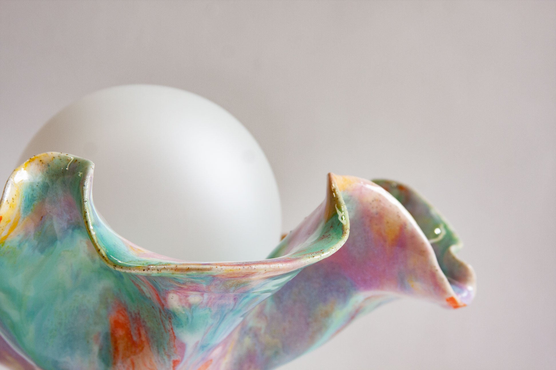 Close-up of the ruffled rim of a handmade ceramic Oyster table lamp curving around the white globe, glazed in green and purple.