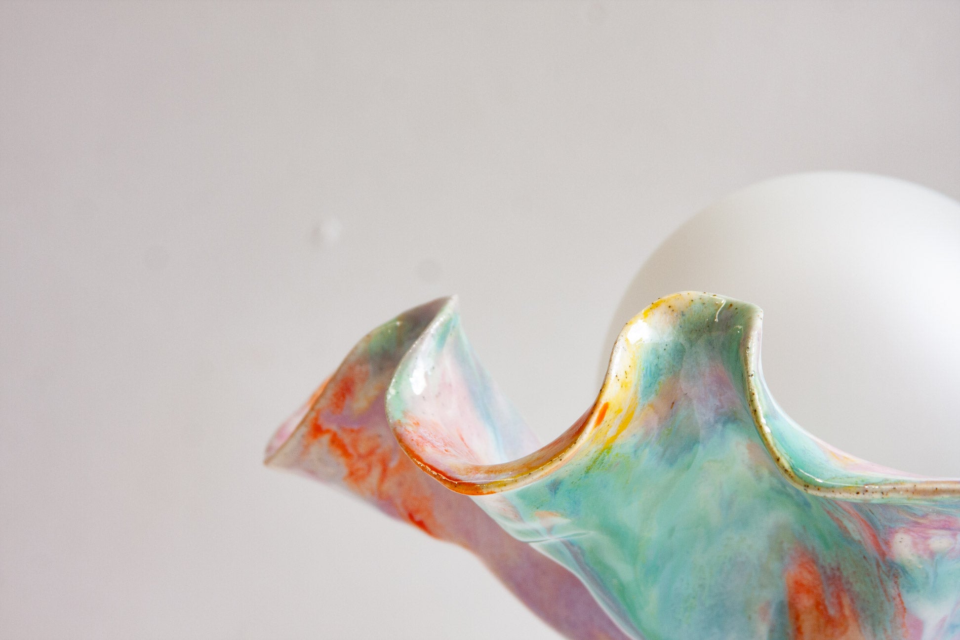 Close-up of the edge of a handmade ceramic Oyster table lamp against the white globe, the rim glazed in green, orange and purple.