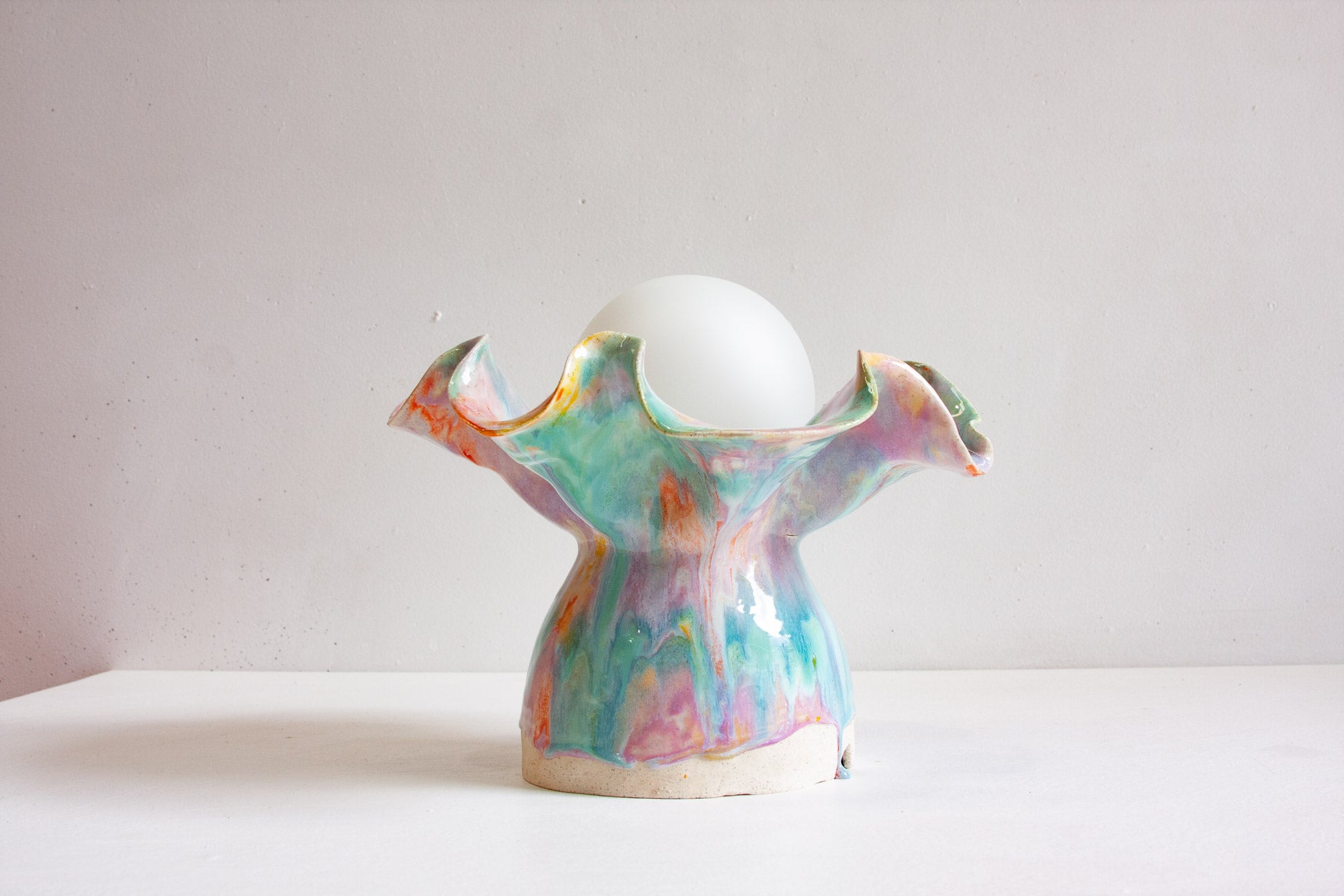 A handmade ceramic Oyster table lamp seen front on, the ruffled base glazed in purple, green and orange below a round white globe.